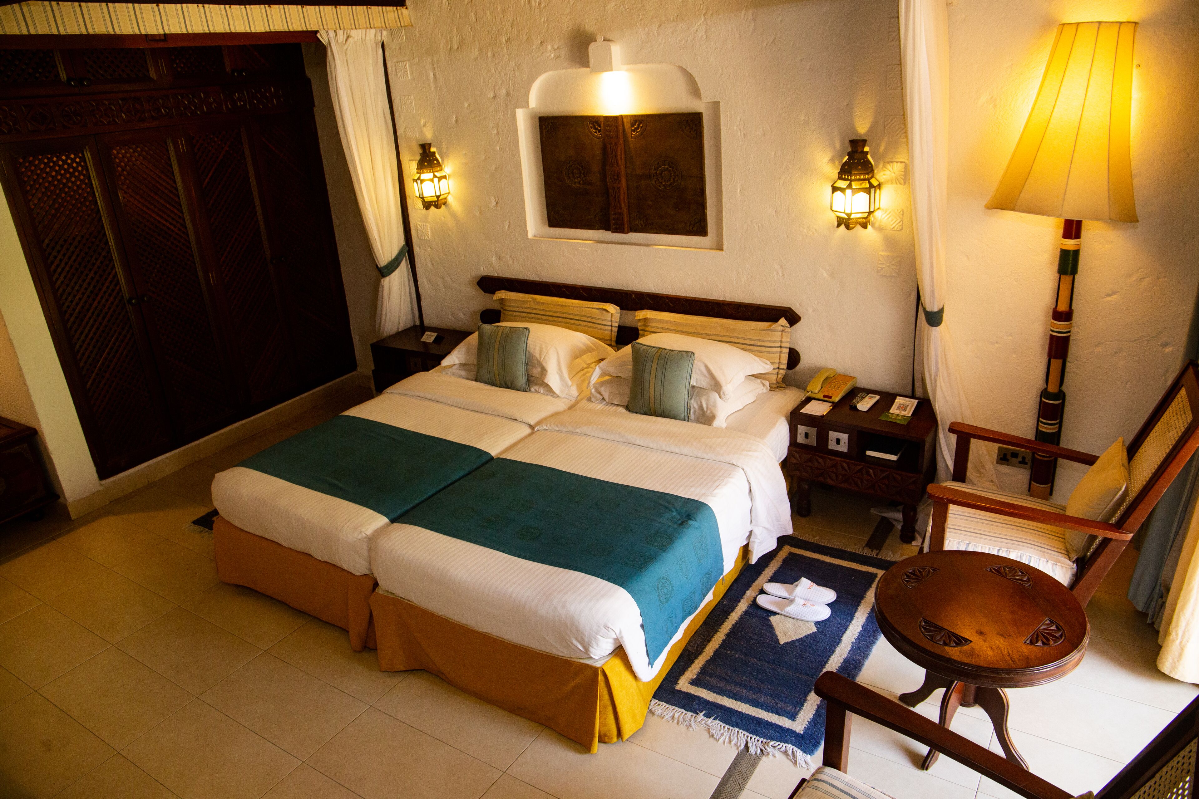 A double bed with a green and white bedspread, two bedside tables with lamps, a wooden chair, and a small rug on the floor.