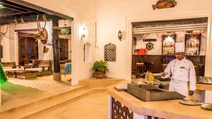 Un chef en la barra con una parrilla, rodeado de un interior rústico con muebles de madera y una planta en maceta.