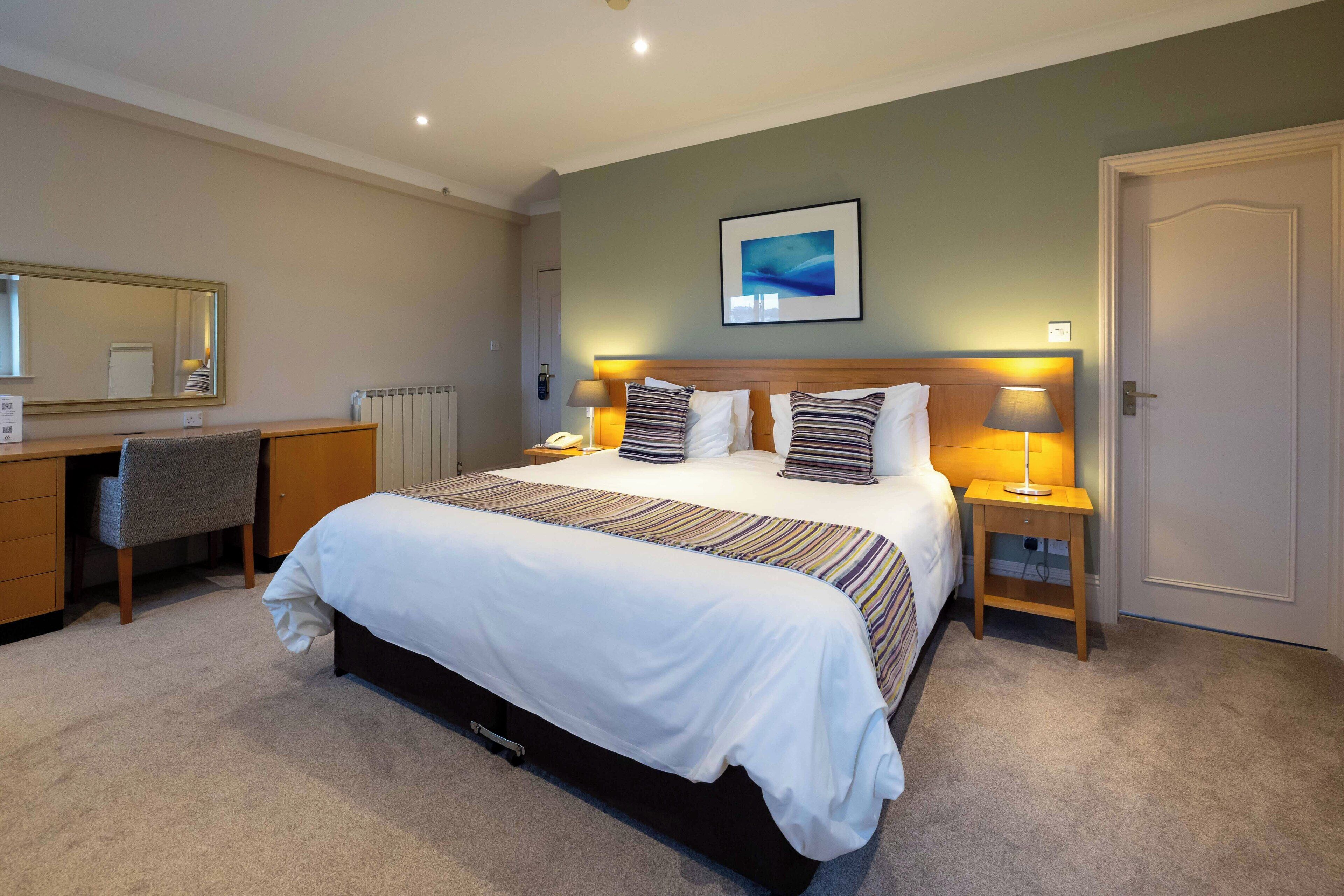 Une chambre d’hôtel avec un grand lit, des tables de chevet avec des lampes, un bureau avec une chaise, un radiateur et un tableau encadré au mur.