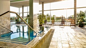 An indoor swimming pool with clear glass walls, stone flooring, and a sunlit area with chairs and potted plants. - Hotel de France (St. Saviour)