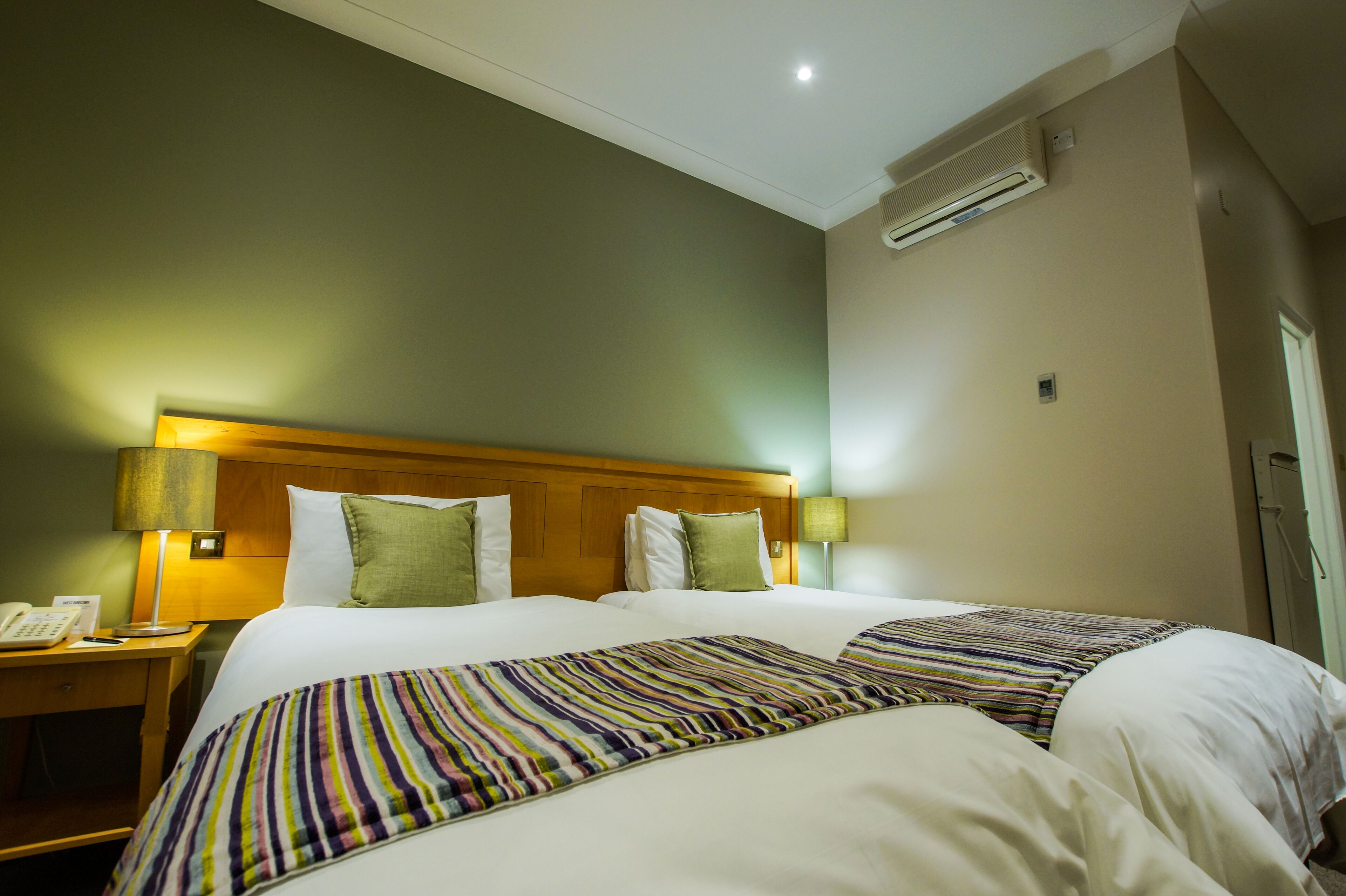 Une chambre d’hôtel avec deux lits, une tête de lit en bois, une table de chevet avec une lampe et un appareil de climatisation fixé au mur.