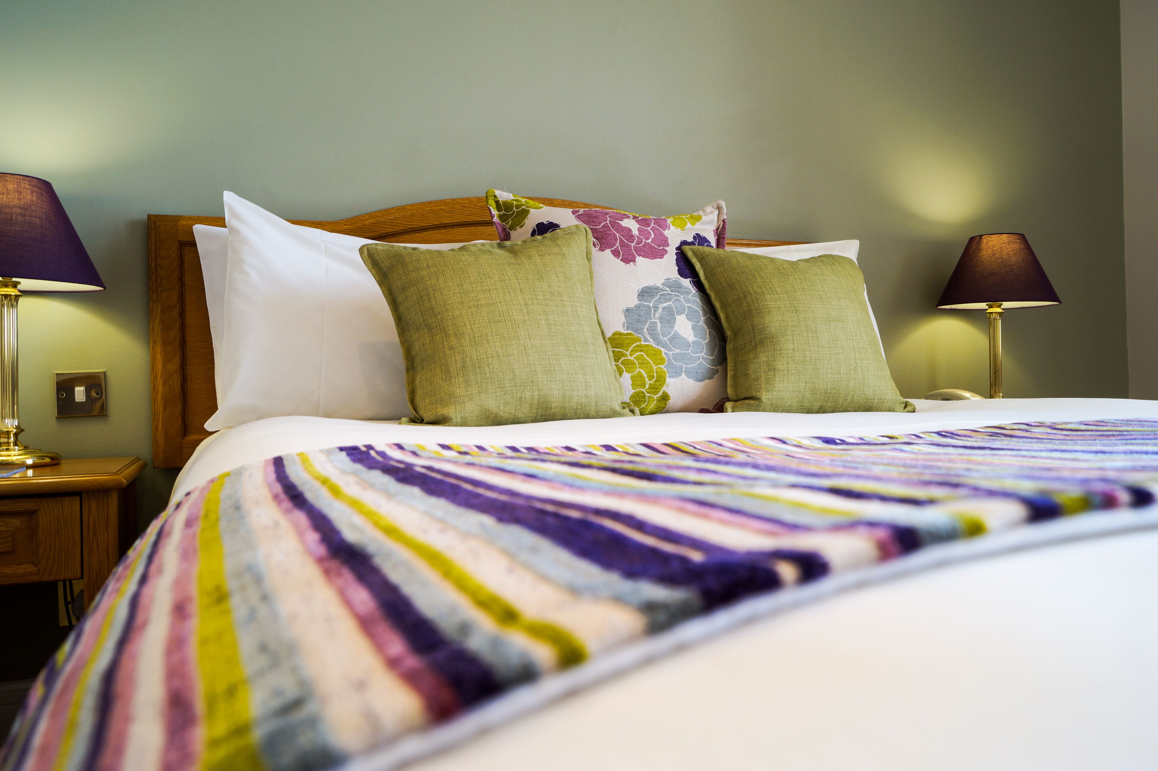 A neatly made bed with a colorful striped blanket, two green pillows, and a floral pillow on a wooden headboard. There are two bedside lamps on either side of the bed.