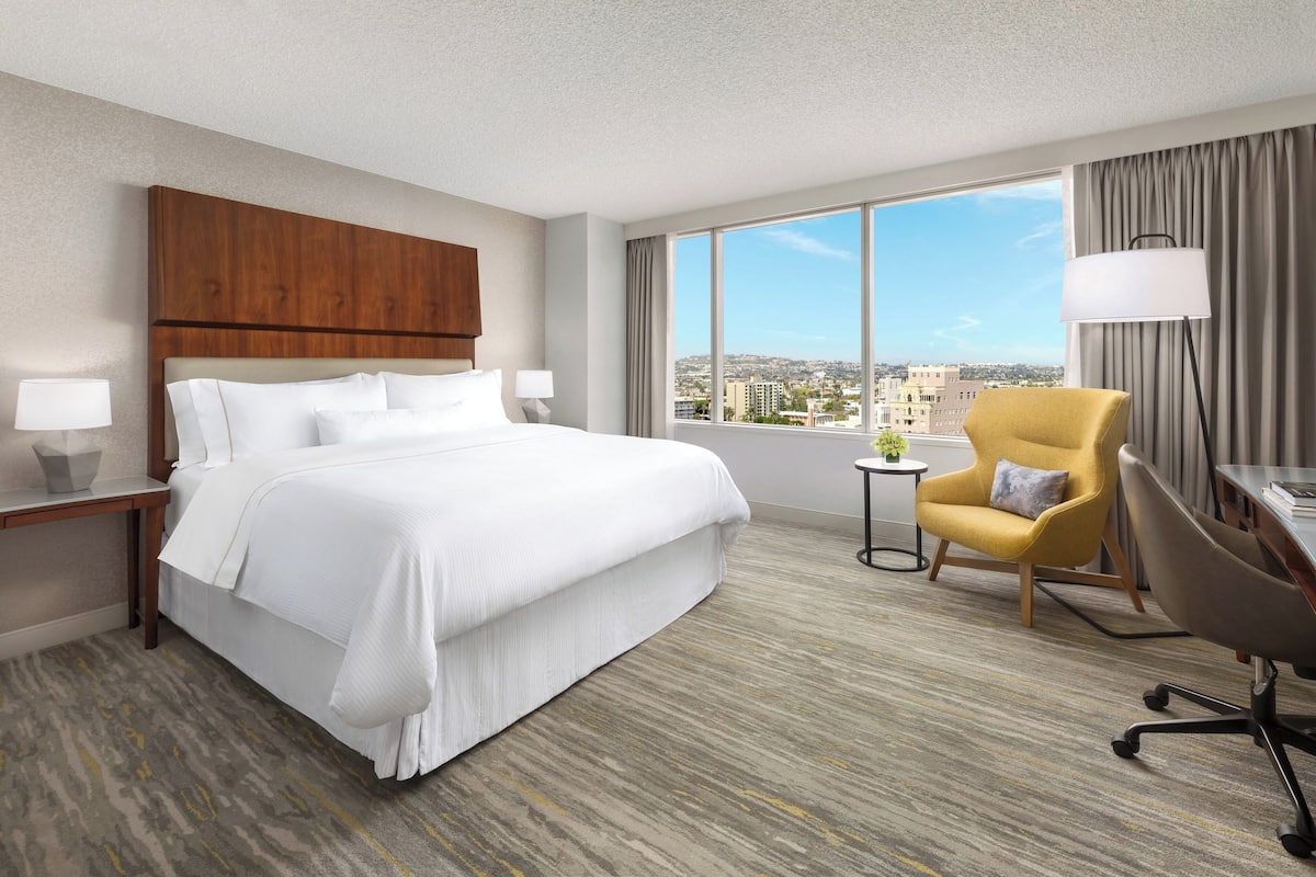 A hotel room with a large bed, a desk with a chair, a small table with a lamp, and a view of the city through large windows.