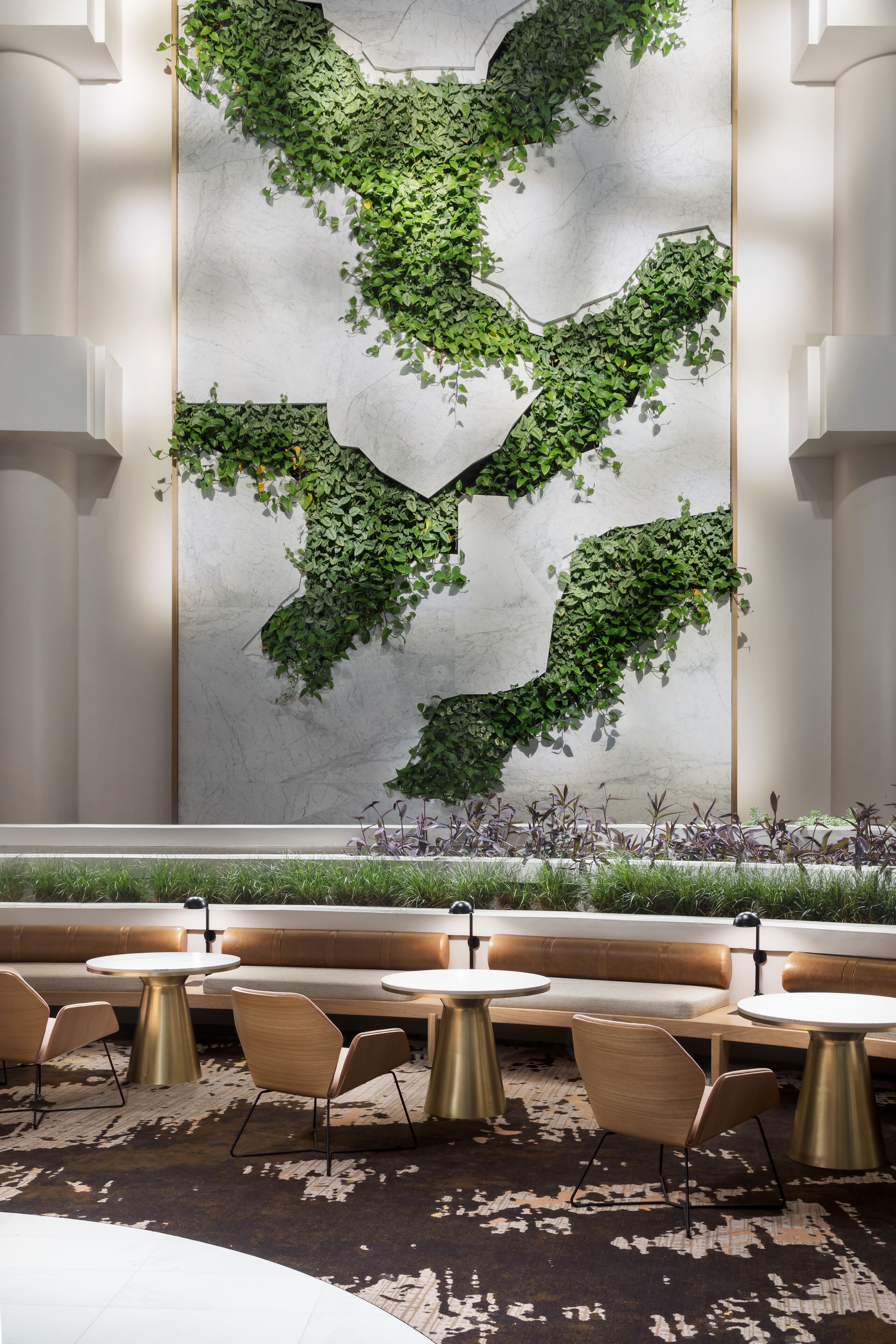 Un interior moderno con un diseño de planta verde montado en la pared, una zona de estar con mesas y sillas redondas, y una alfombra estampada.