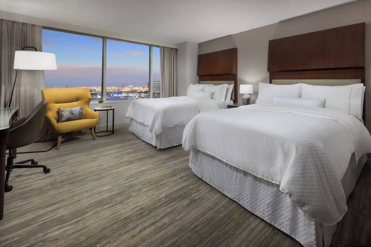 A hotel room with two beds, a desk, a chair, and a large window offering a city view.