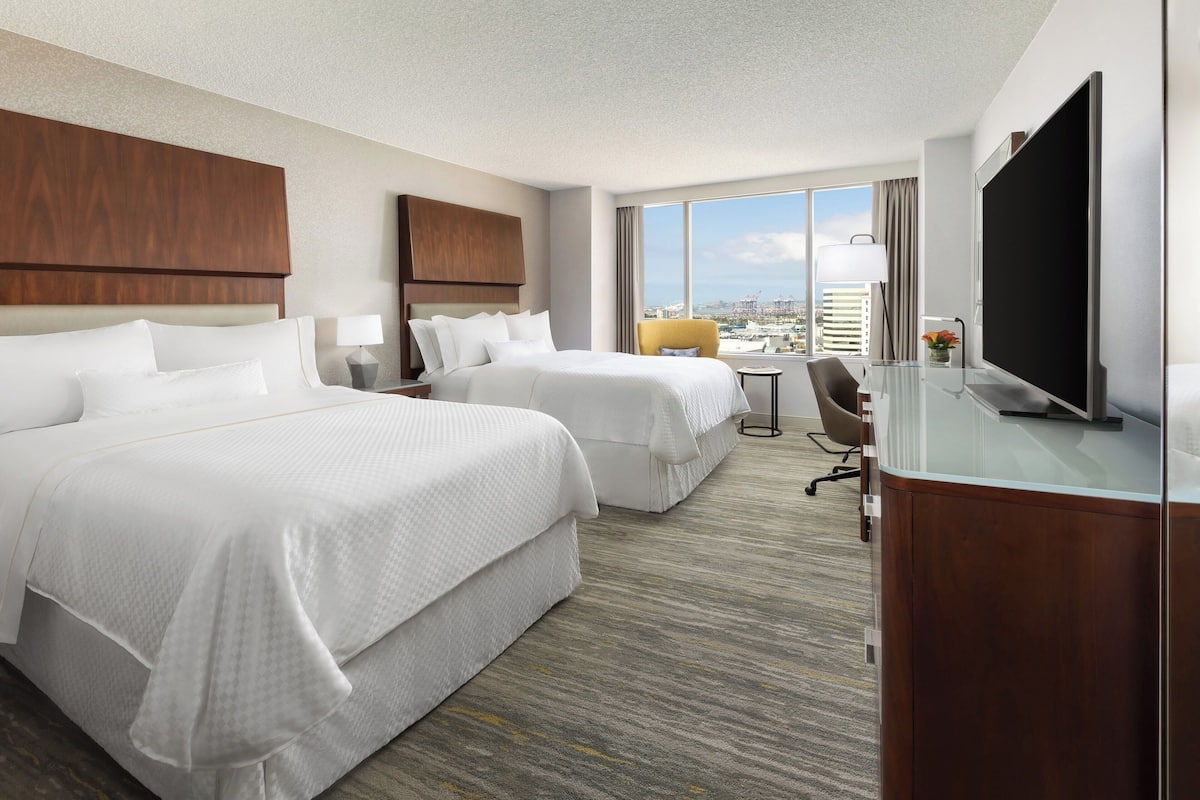 A hotel room with two beds, a TV, a desk, and a view of the city.