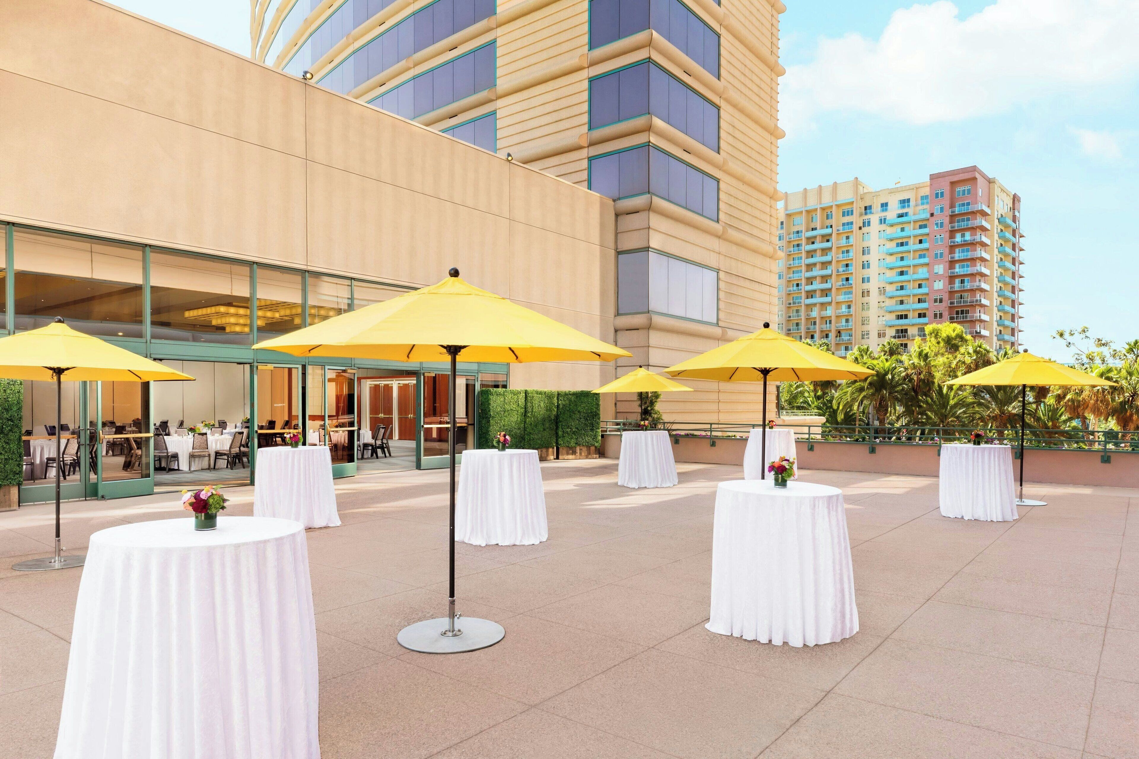 Un patio de hotel con mesas redondas cubiertas con manteles blancos y sombrillas amarillas, frente a la fachada de un edificio moderno.
