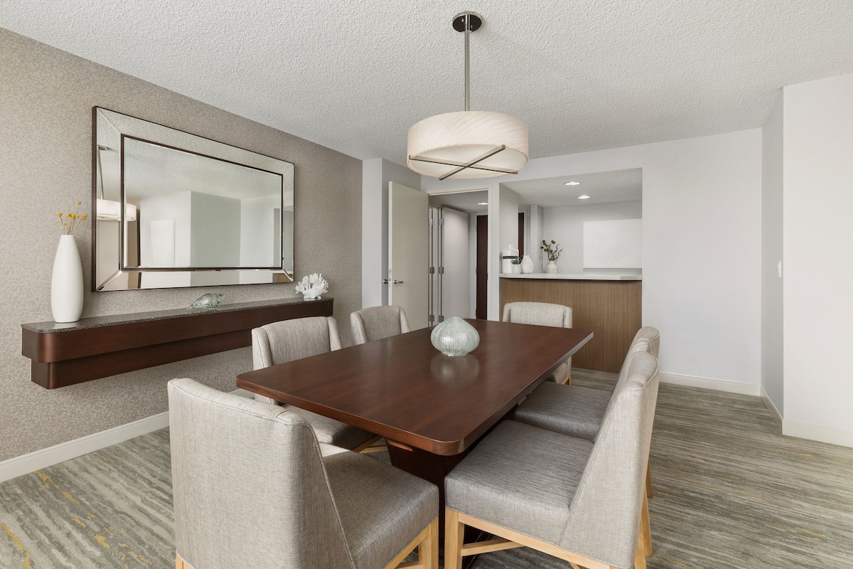 A modern dining area with a rectangular table, six upholstered chairs, a large mirror, and a hanging light fixture.