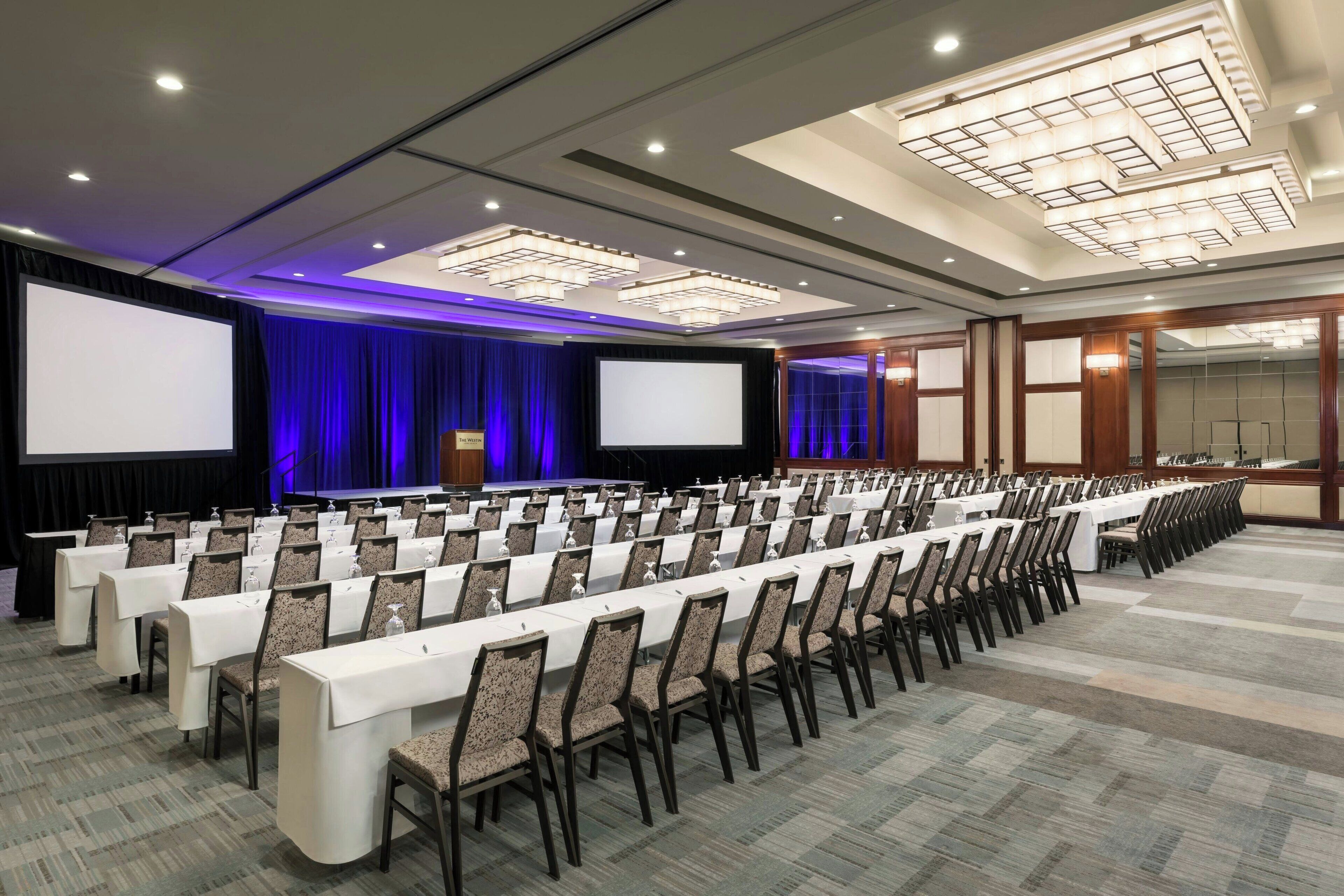 Un salón de conferencias con filas de mesas y sillas, pantallas grandes y un escenario con cortinas.