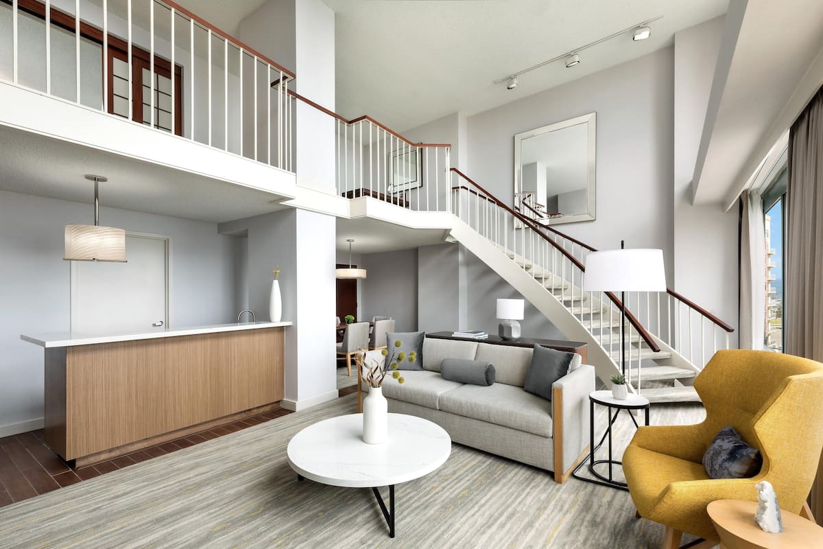 A spacious living area with a high ceiling, a staircase, a sofa, a yellow armchair, and a round coffee table.