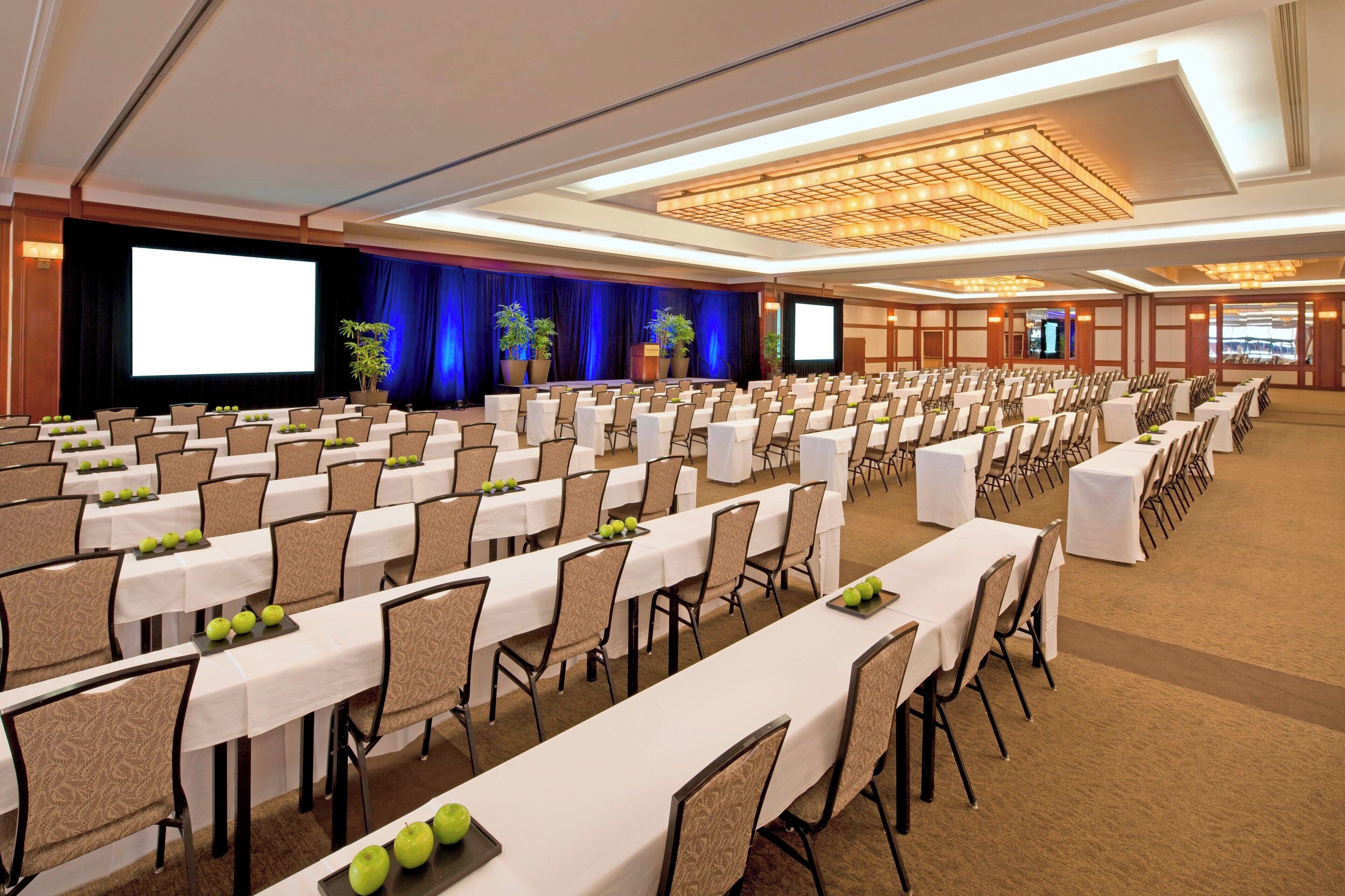 Une grande salle de conférence avec des rangées de tables et de chaises, une scène dotée d’un grand écran et des plantes en pot.