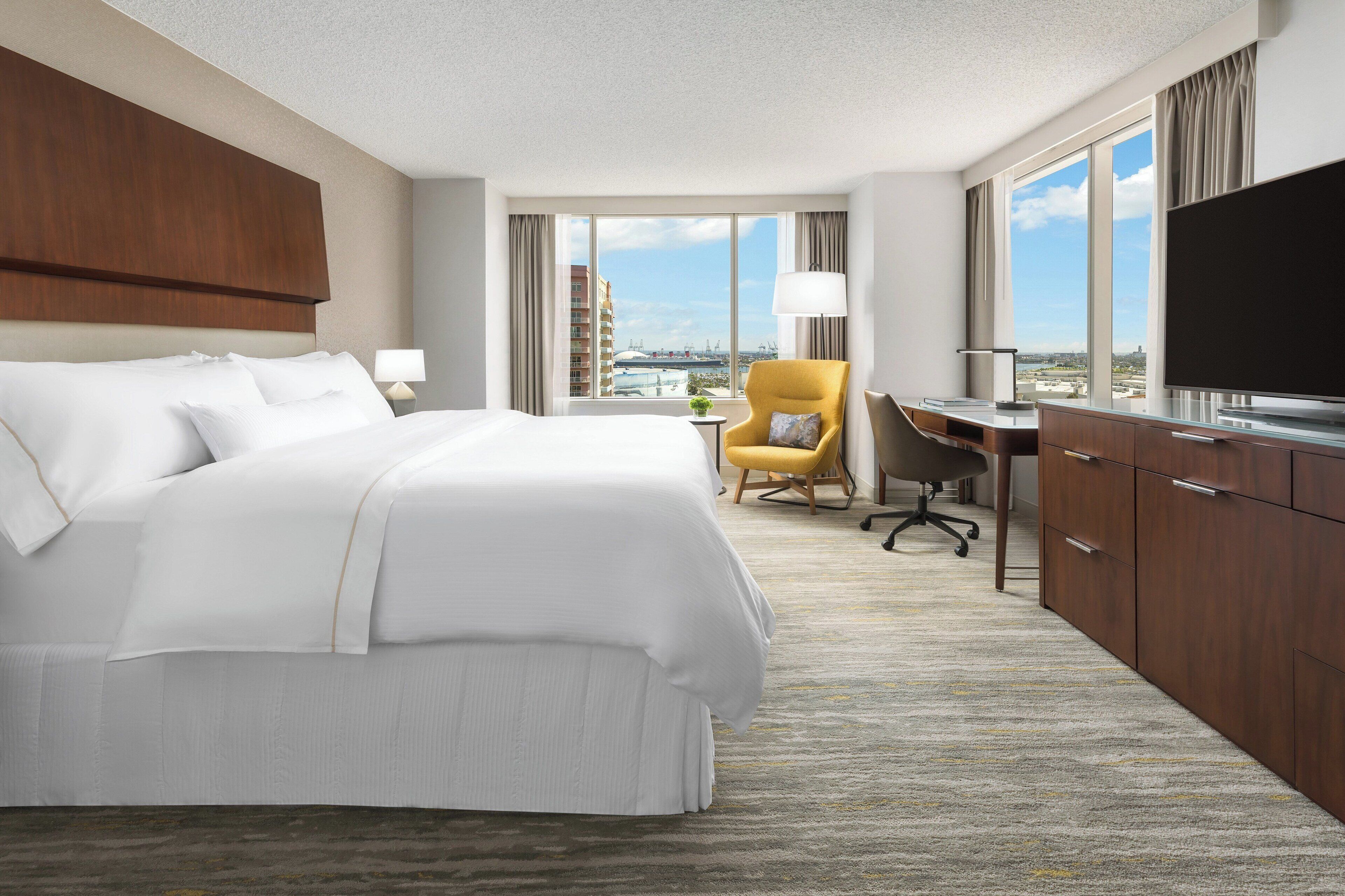 A hotel room with a large bed, a desk, a chair, a TV, and a view of the city.