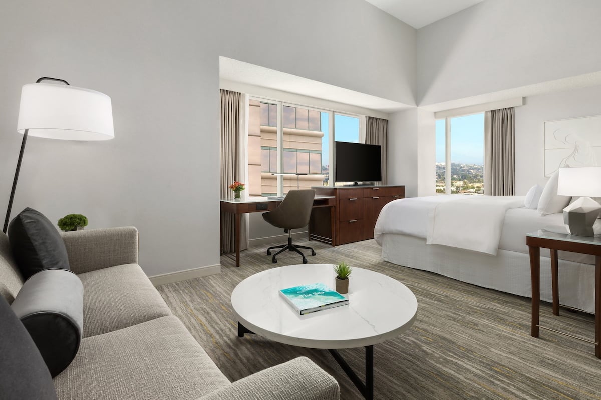 A hotel room with a sofa, a round coffee table with a book, a desk with a chair, a TV, and a bed.