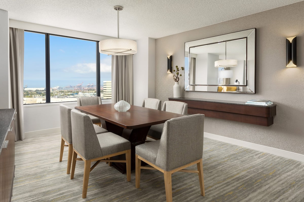 A modern hotel room with a large dining table, comfortable chairs, a large mirror, and a view of the cityscape.