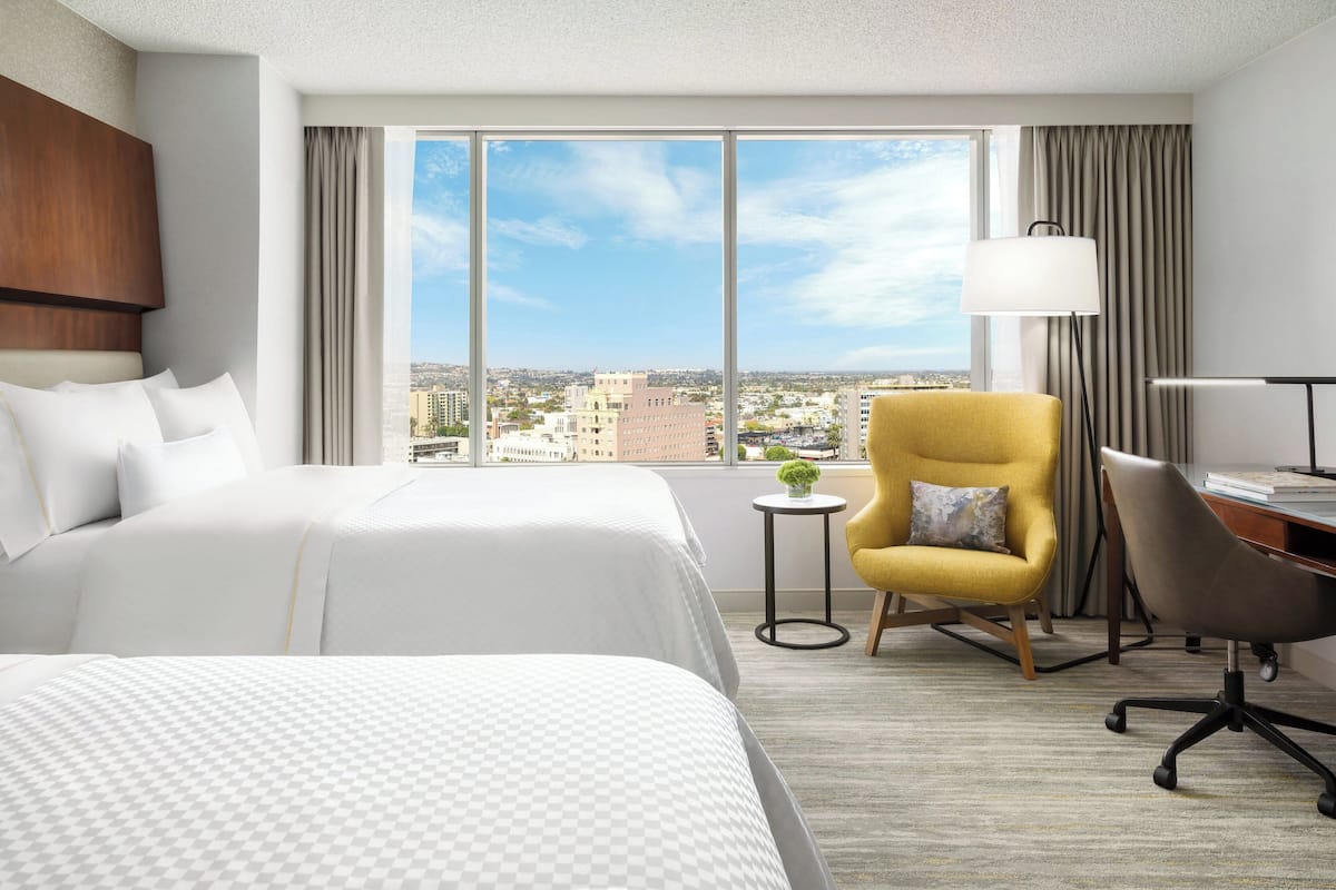 A hotel room with two beds, a desk, a chair, a lamp, and a view of the city.