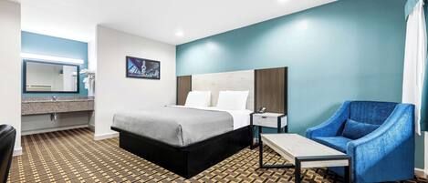 Une chambre d’hôtel comprenant un lit, une chaise, une petite table, une table de chevet, un miroir et une salle de bain avec un lavabo et un porte-serviettes.