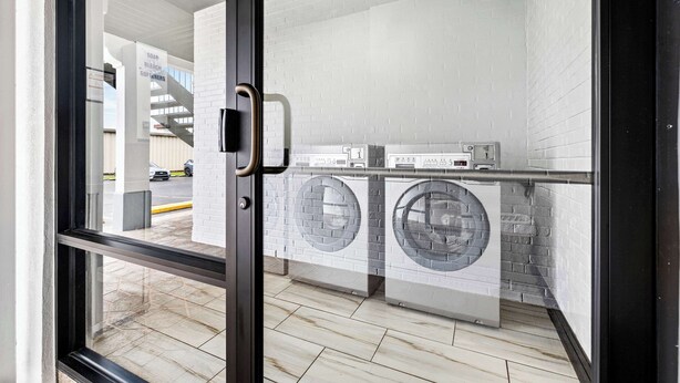 Une buanderie moderne équipée de deux machines à laver à chargement frontal et d’un sèche-linge, avec des murs en briques blanches et une porte en verre donnant sur l’extérieur.