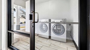 A modern laundry room with two front-loading washing machines and a dryer, white brick walls, and a glass door leading outside.