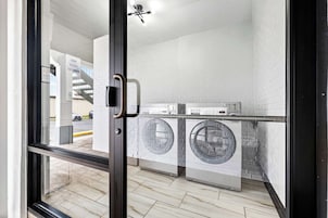 A modern laundry room with two front-loading washing machines and a dryer, white brick walls, and a glass door leading outside.