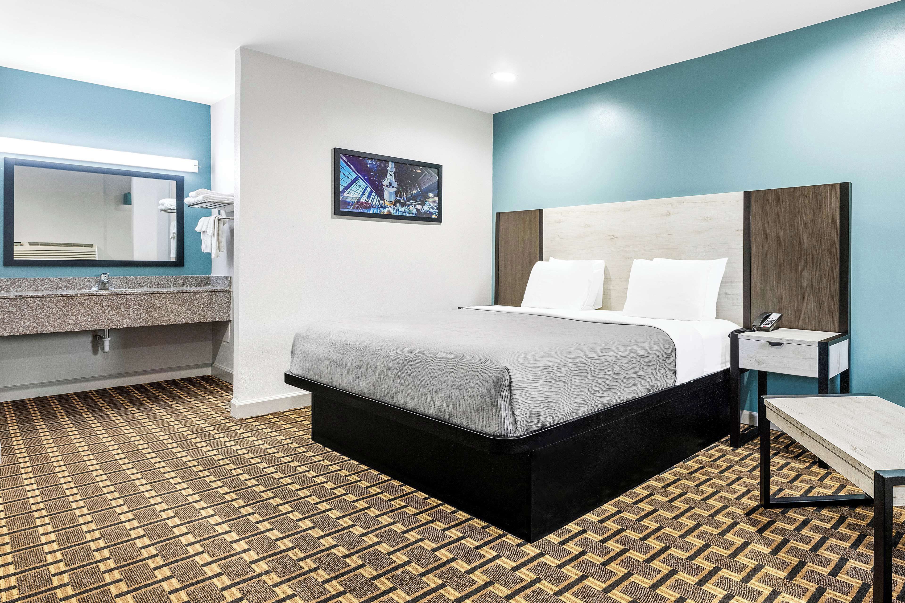 Une chambre d’hôtel comprenant un lit, une table de chevet, un bureau, une salle de bain avec un lavabo et un miroir, et un sol à motifs.