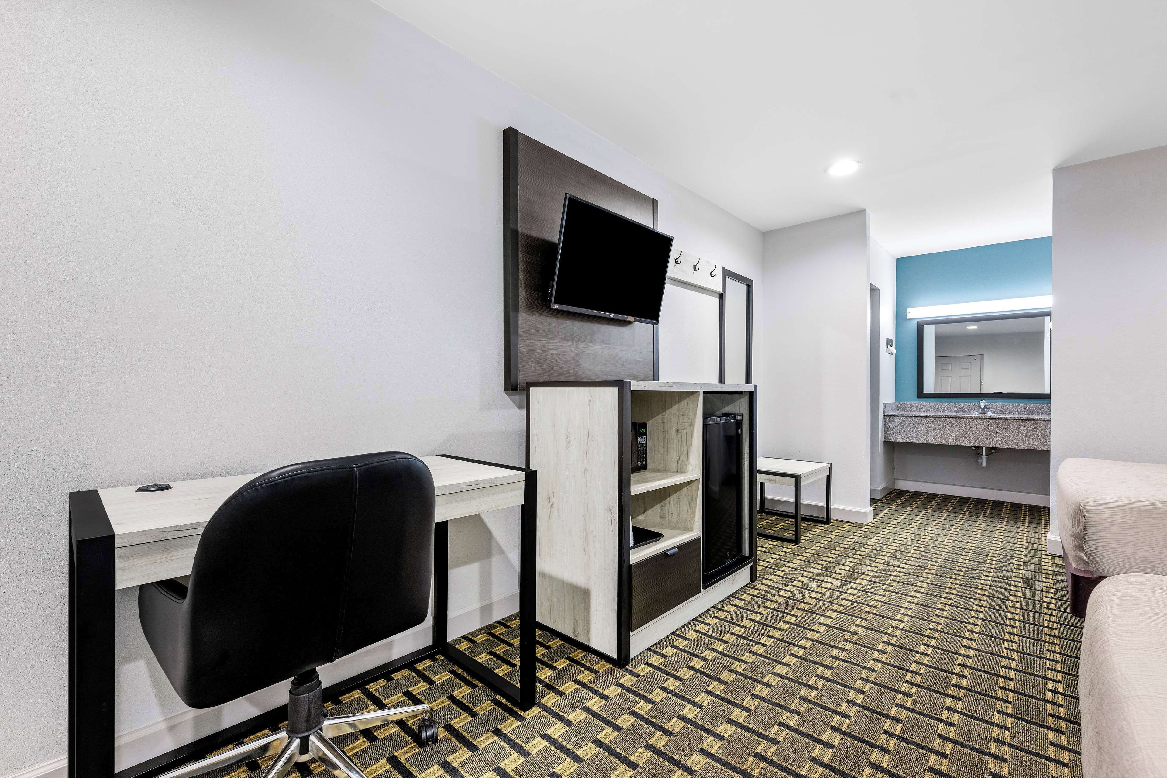 A hotel room with a desk, chair, TV, mini-fridge, and a bed.
