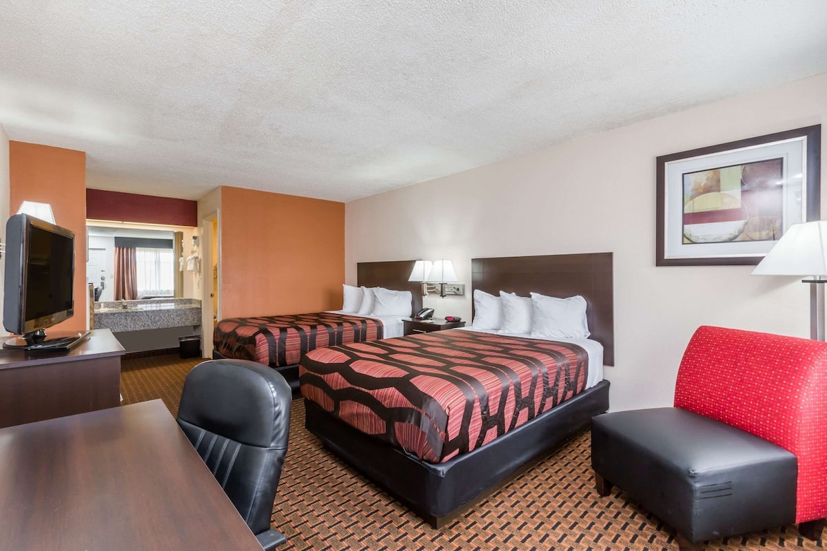 A hotel room with two beds, a desk with a television, a red chair, and a framed picture on the wall.