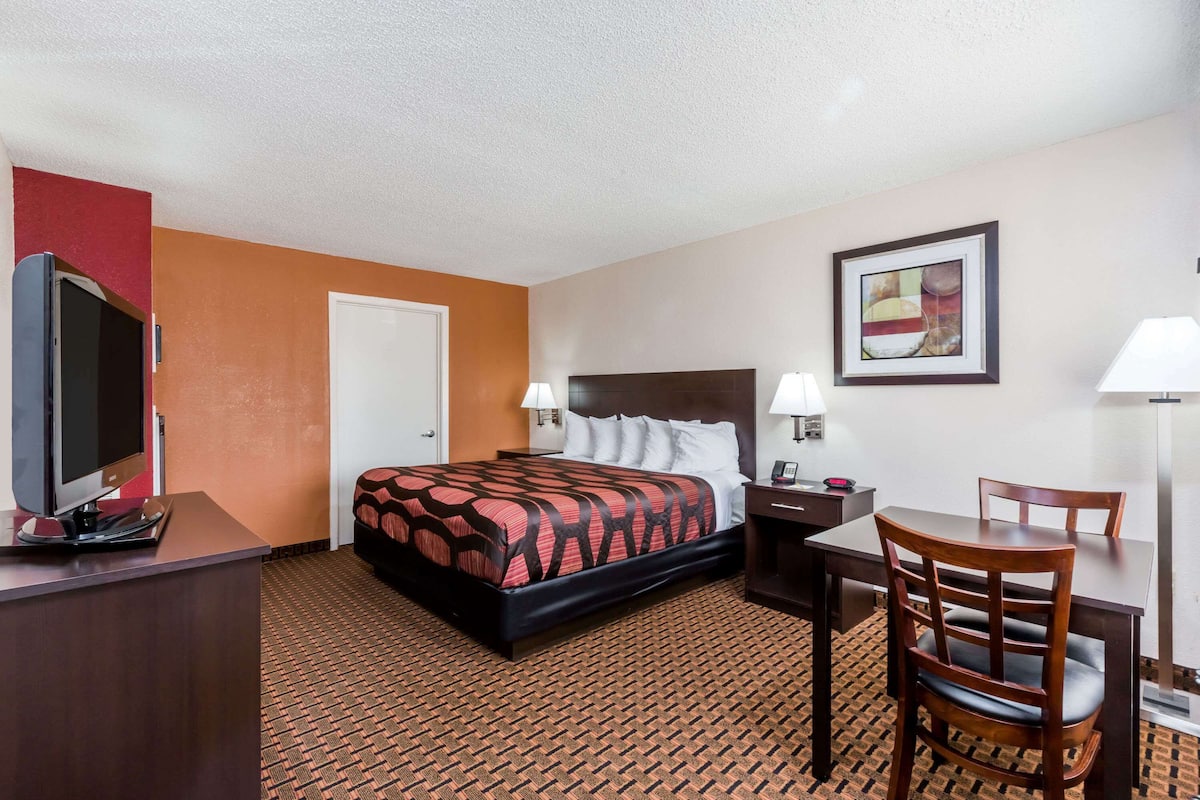 A hotel room with a bed, desk, chair, TV, and artwork on the wall.