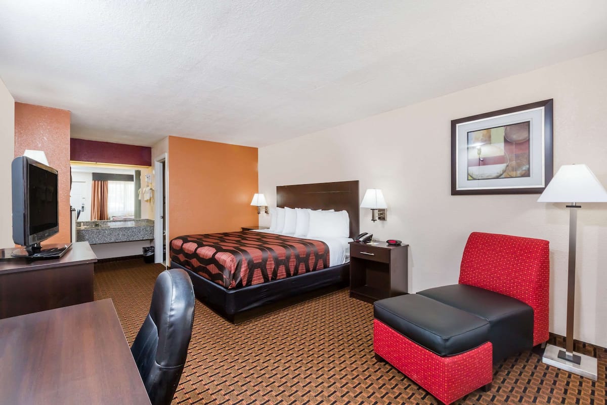 A hotel room with a bed, a desk with a television, a red chair, and a lamp.
