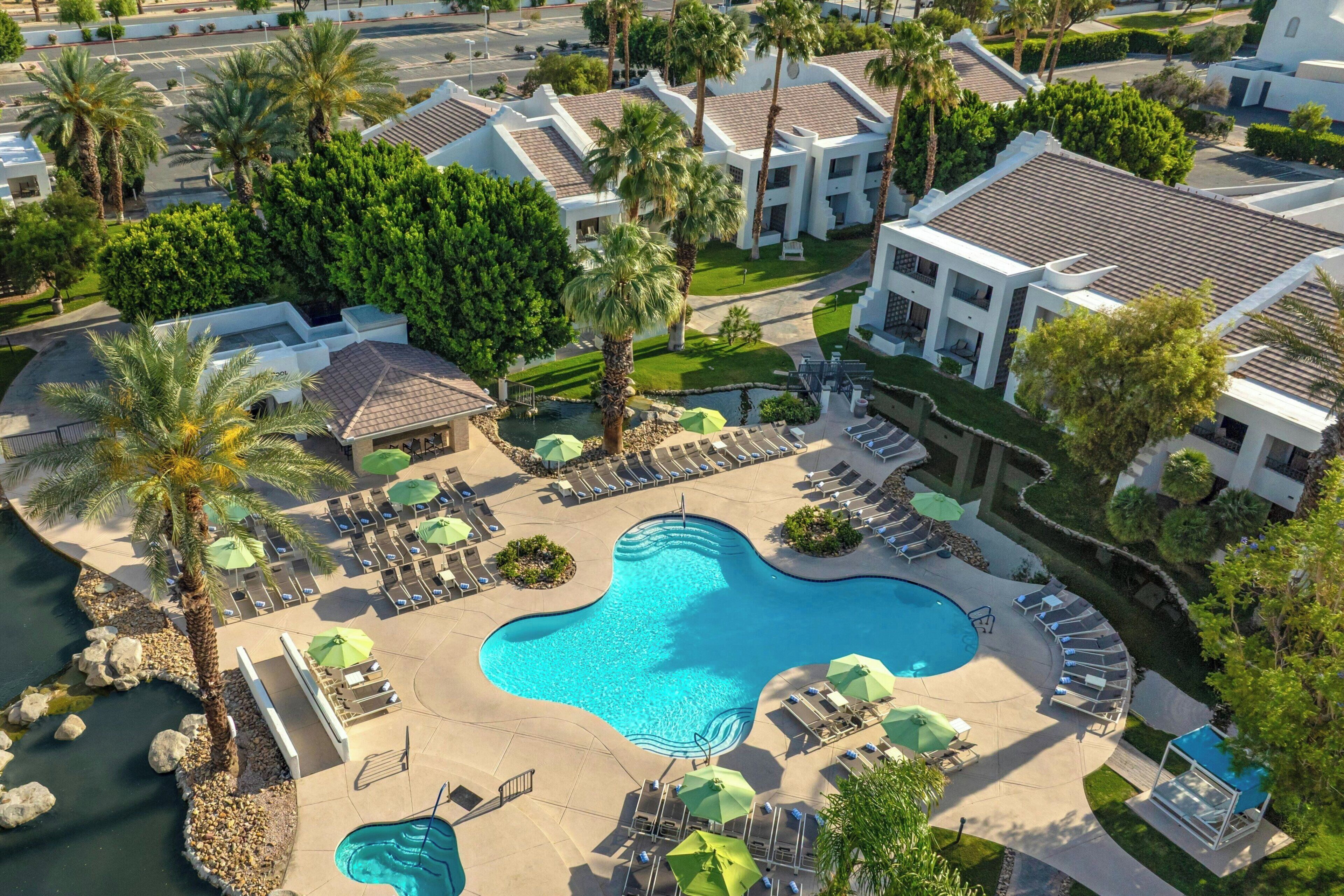 A resort with a swimming pool, lounge chairs, palm trees, and a parking area.