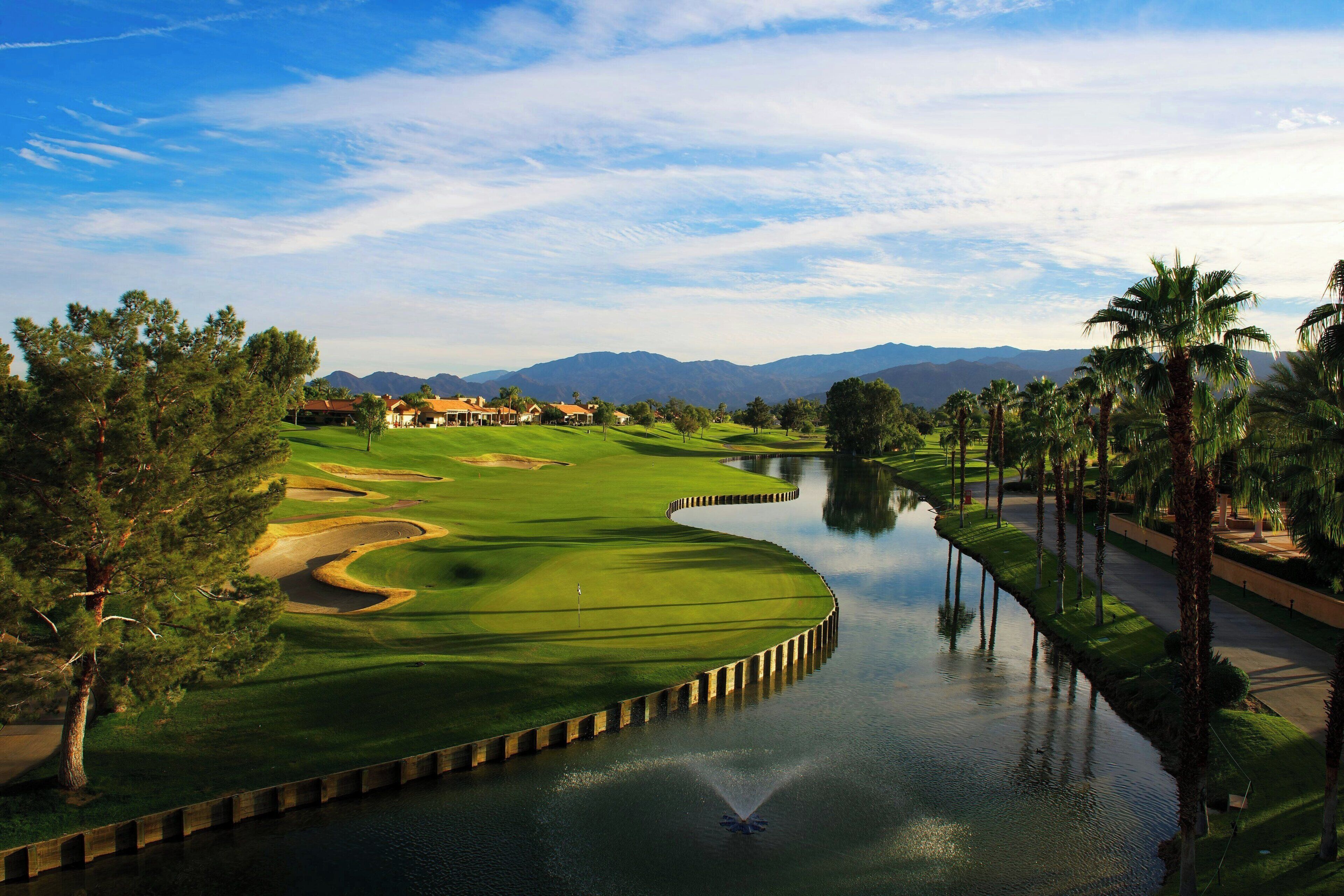 Ein Golfplatz mit einem Wasserspiel, Palmen und Bergen im Hintergrund.