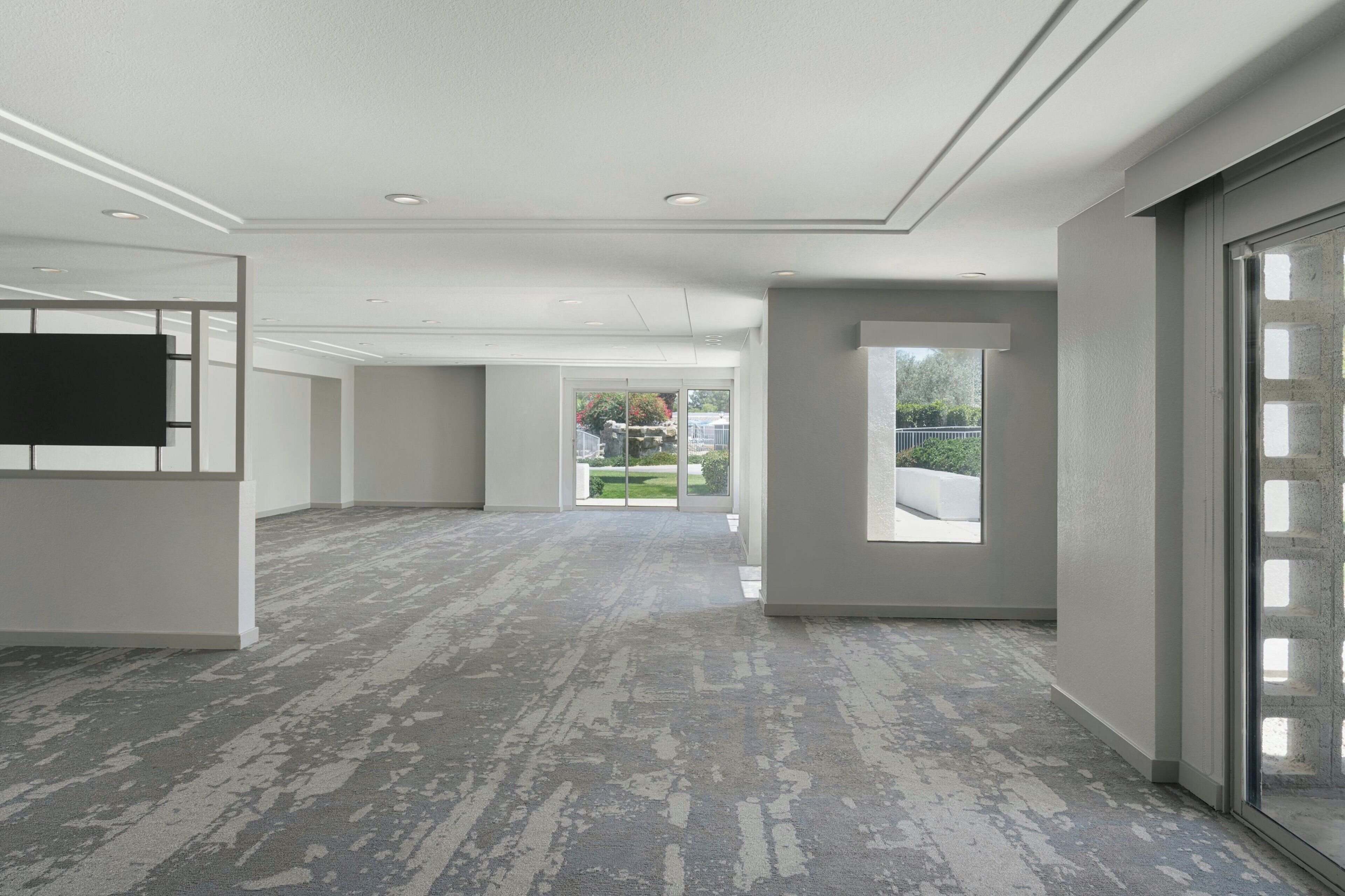 A spacious hallway with a grey carpet, large windows, and a flat-screen TV mounted on the wall.