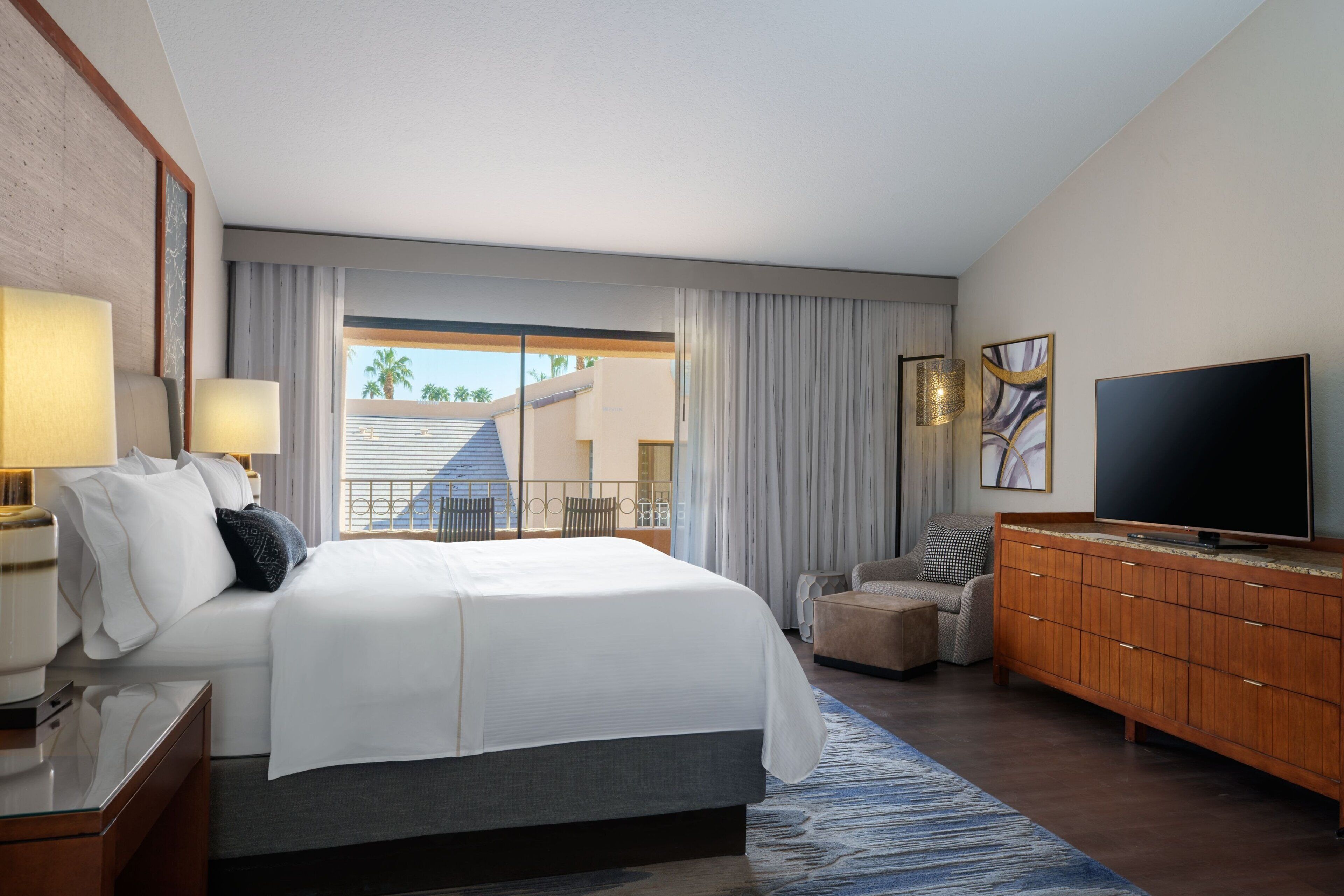 A hotel room with a large bed, a TV, a wooden dresser, and a view of palm trees through the window.