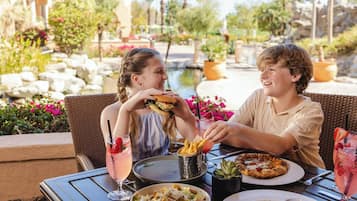 Zwei Personen speisen an einem Tisch im Freien, mit verschiedenen Gerichten, Getränken und Blick auf einen schönen Garten im Hintergrund.