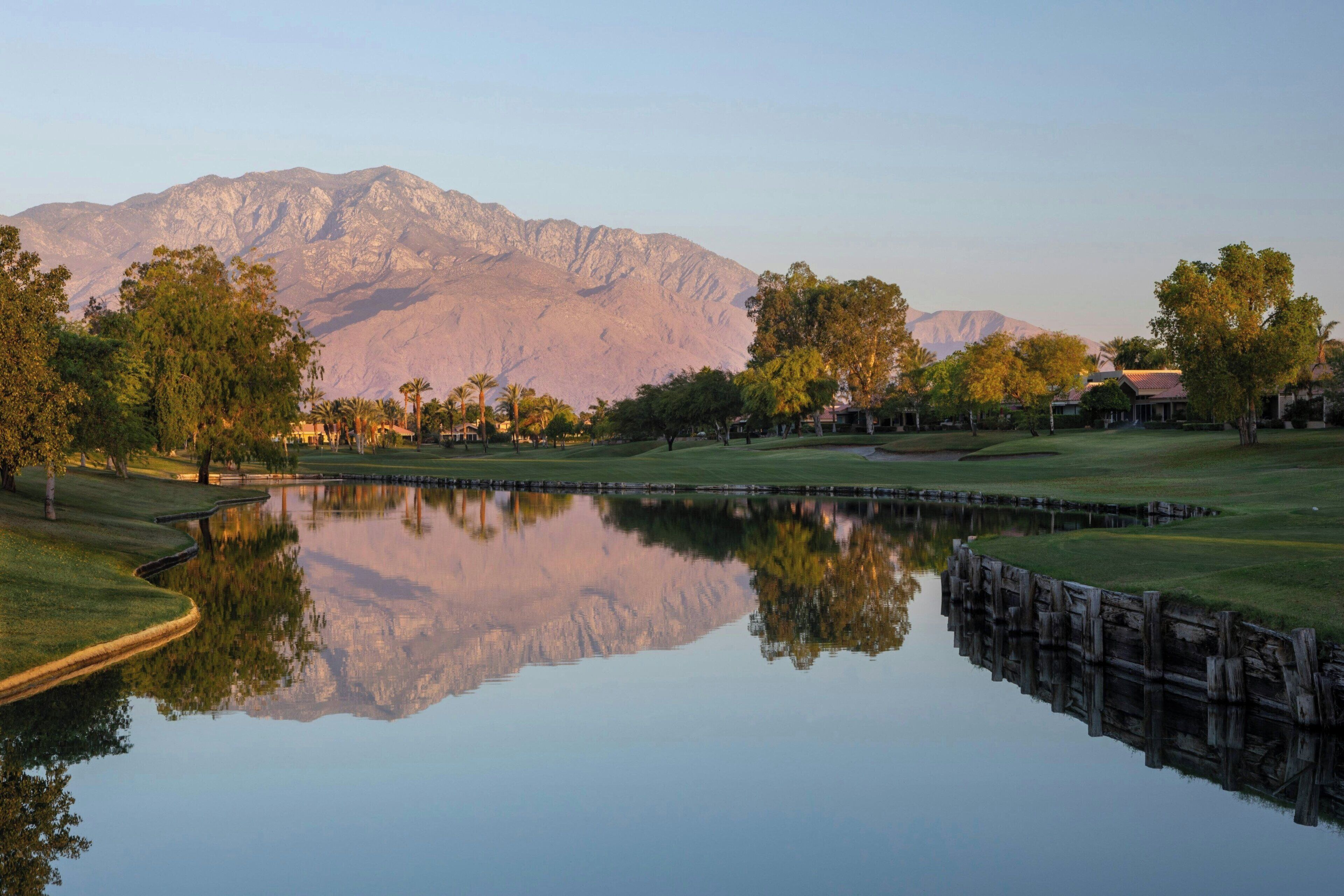 Un campo de golf tranquilo con un estanque reflejante, abundante vegetación y montañas al fondo.