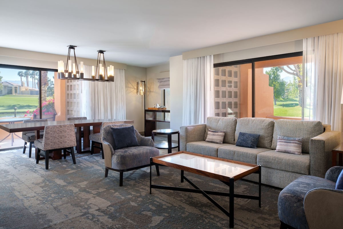 Een ruime woonkamer met een grote bank, een salontafel en een eettafel met stoelen. Een kroonluchter hangt aan het plafond en er is uitzicht op een golfbaan door de ramen.