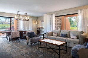Un vaste espace de vie comprenant un grand canapé, une table basse et une table à manger avec des chaises. Un lustre est suspendu au plafond et les fenêtres offrent une vue sur un terrain de golf.