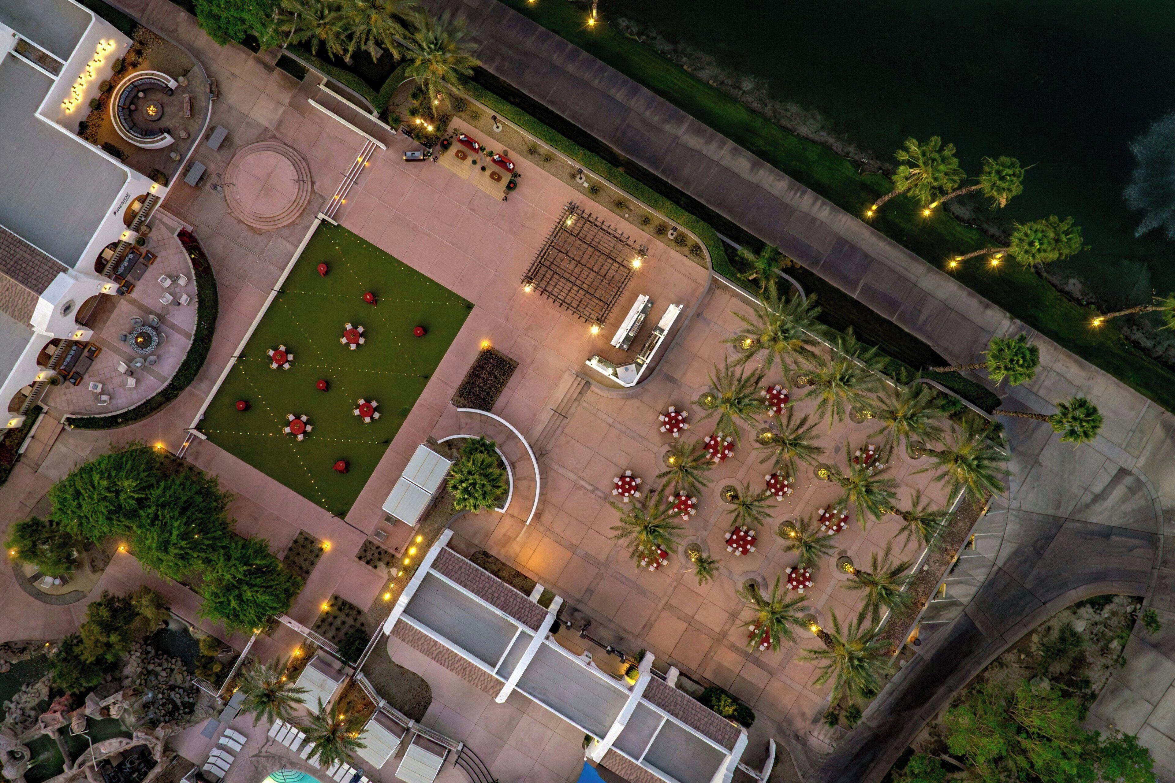 Vue aérienne d’une cour d’hôtel avec une piscine, des espaces de détente en plein air, des palmiers et un pont.