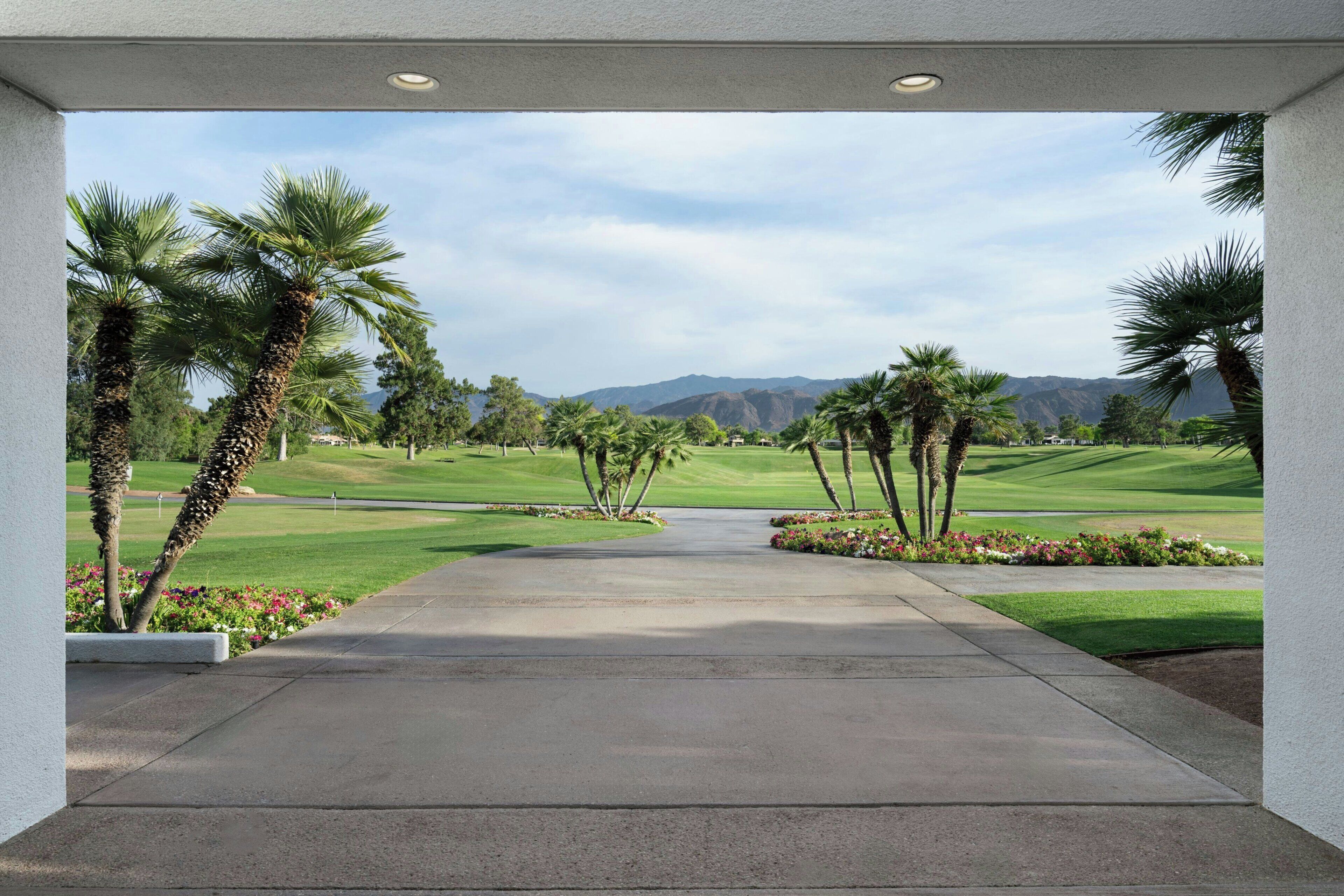 A paved pathway leading to a golf course with palm trees and mountains in the background.