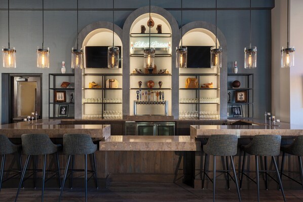 Un espace bar doté d’un long comptoir, de tabourets de bar et d’étagères où sont disposés des verres et des bouteilles.