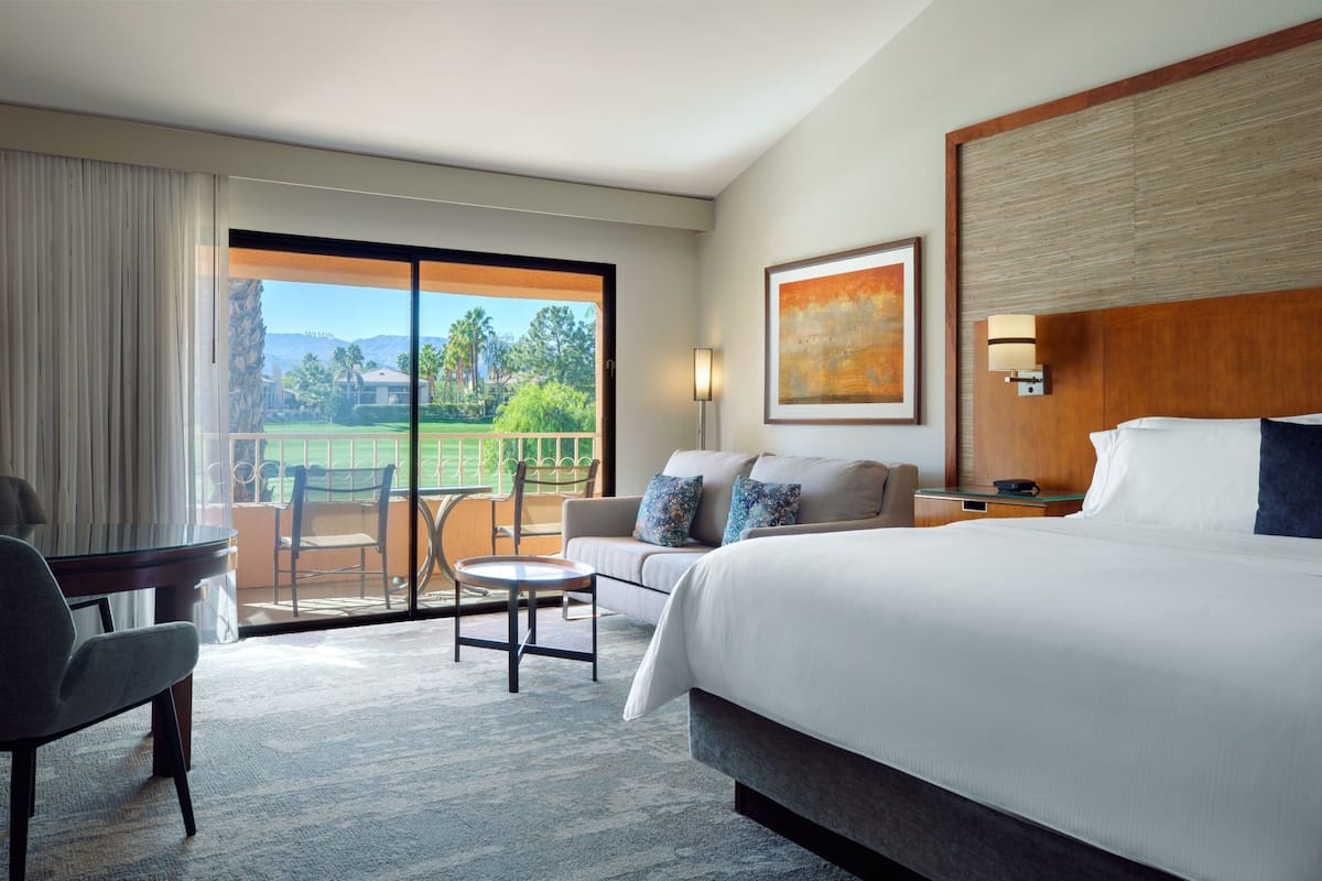 Hotelkamer met een groot bed, een bank, een eettafel en een stoel. Groot raam met uitzicht op een golfbaan en bergen.
