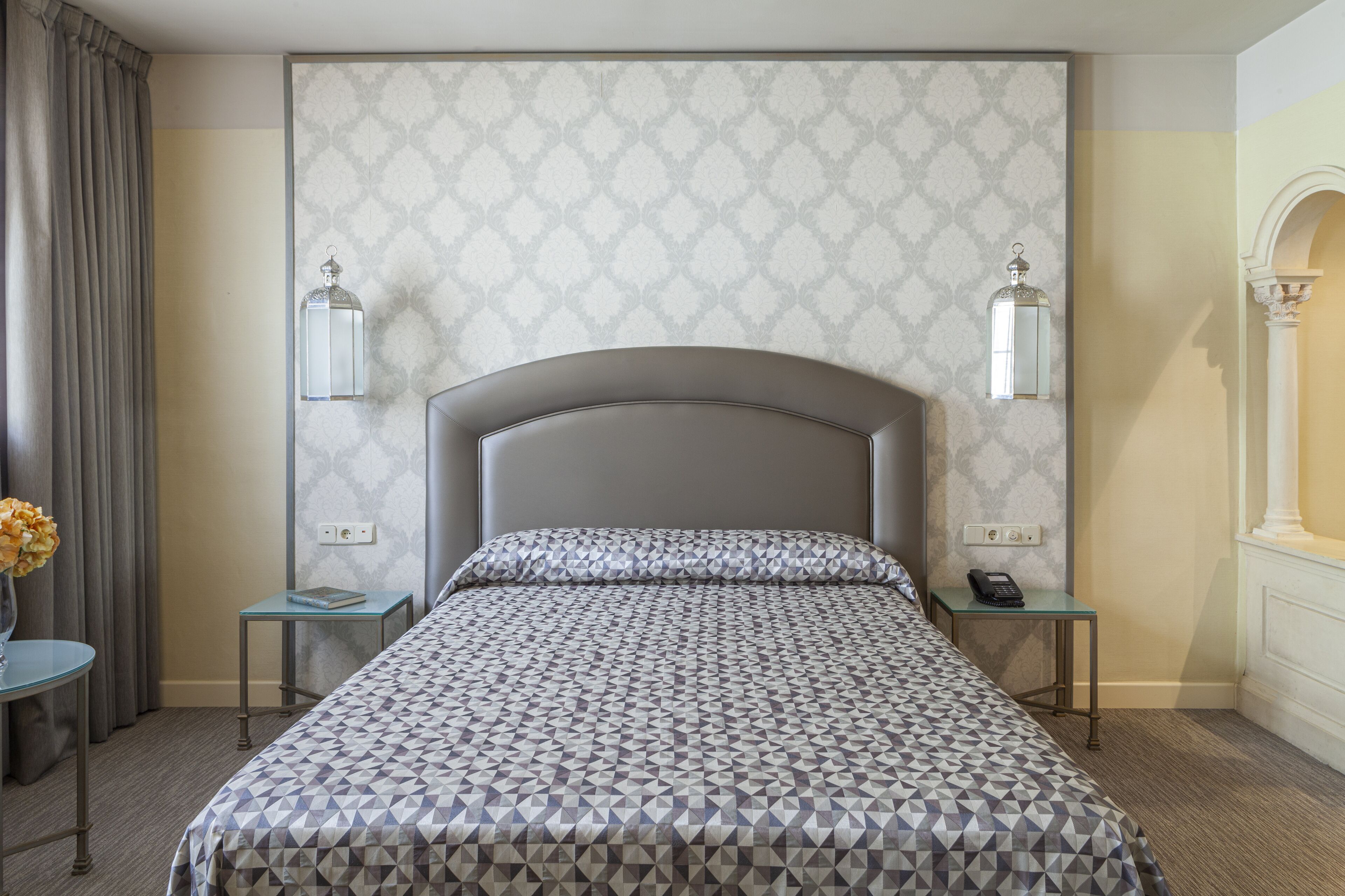 A hotel room with a patterned bedspread, two bedside tables with lamps, a vase with flowers, and a mirror on the wall.