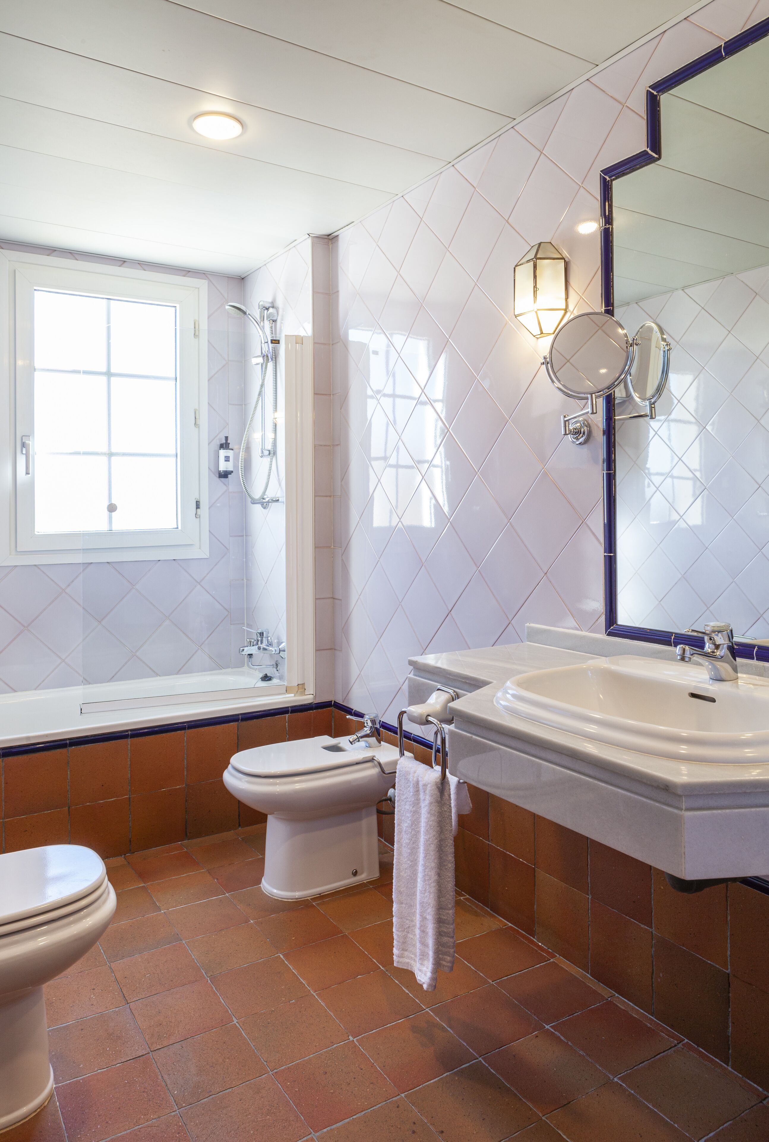 Un baño con bañera, inodoro y lavamanos, con ventana, paredes de azulejos y un espejo.