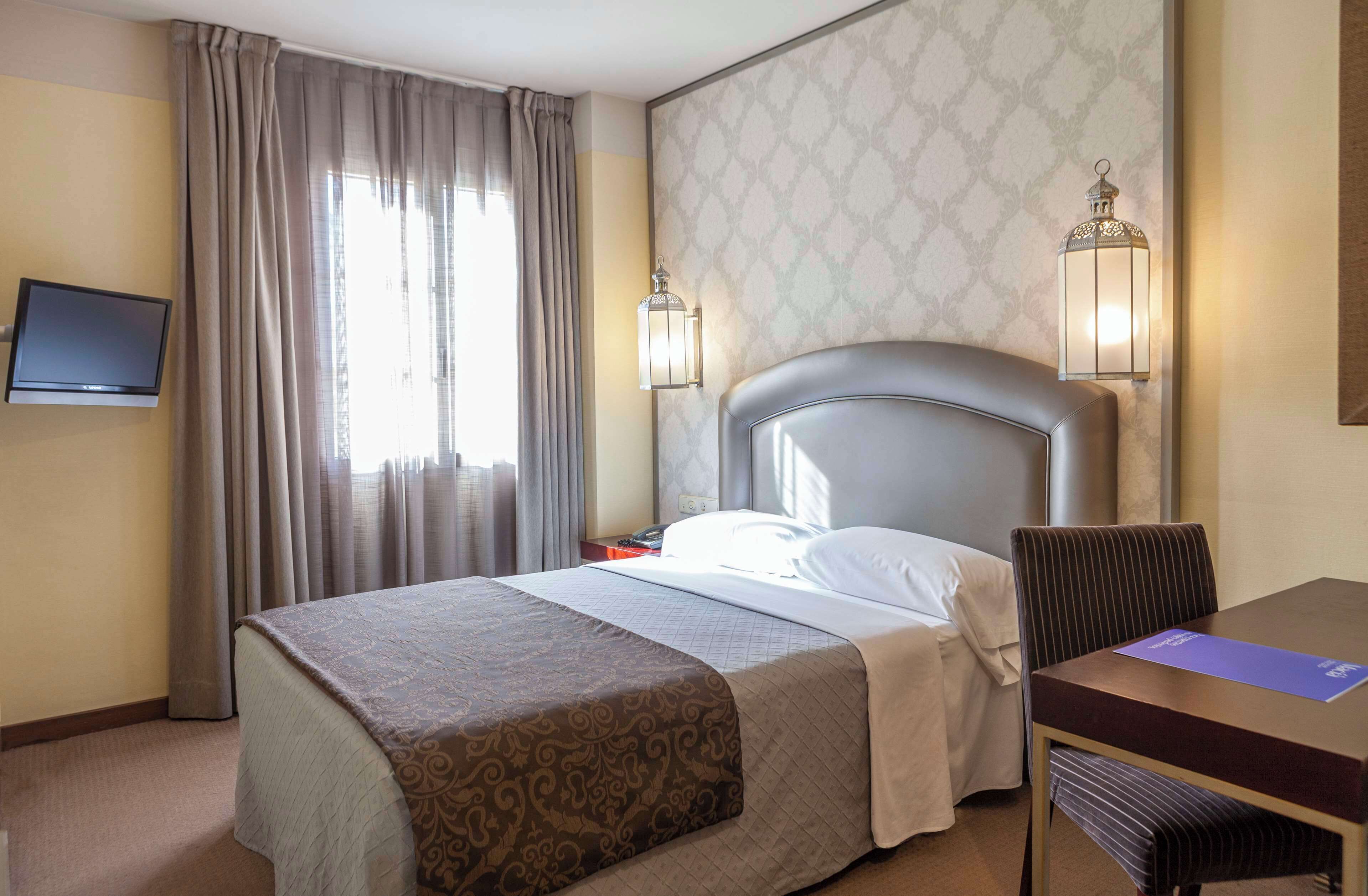 Habitación de hotel con una cama grande, un escritorio, una silla, un televisor y dos lámparas fijadas a la pared.