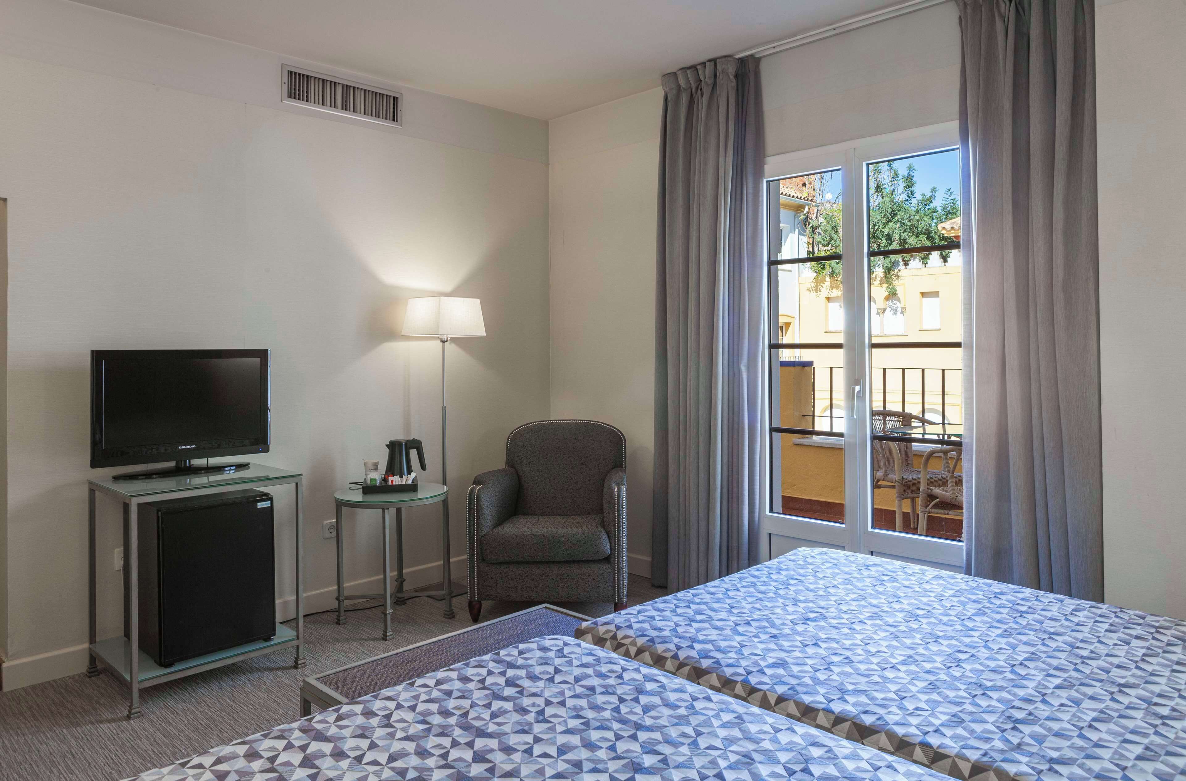 A hotel room with two beds, a TV, a small fridge, a lamp, and a chair.