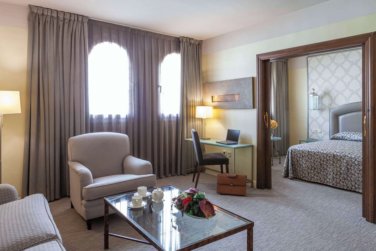 Una habitación de hotel con cama, escritorio con ordenador portátil, silla, sofá y mesa de centro con tazas y un arreglo floral.