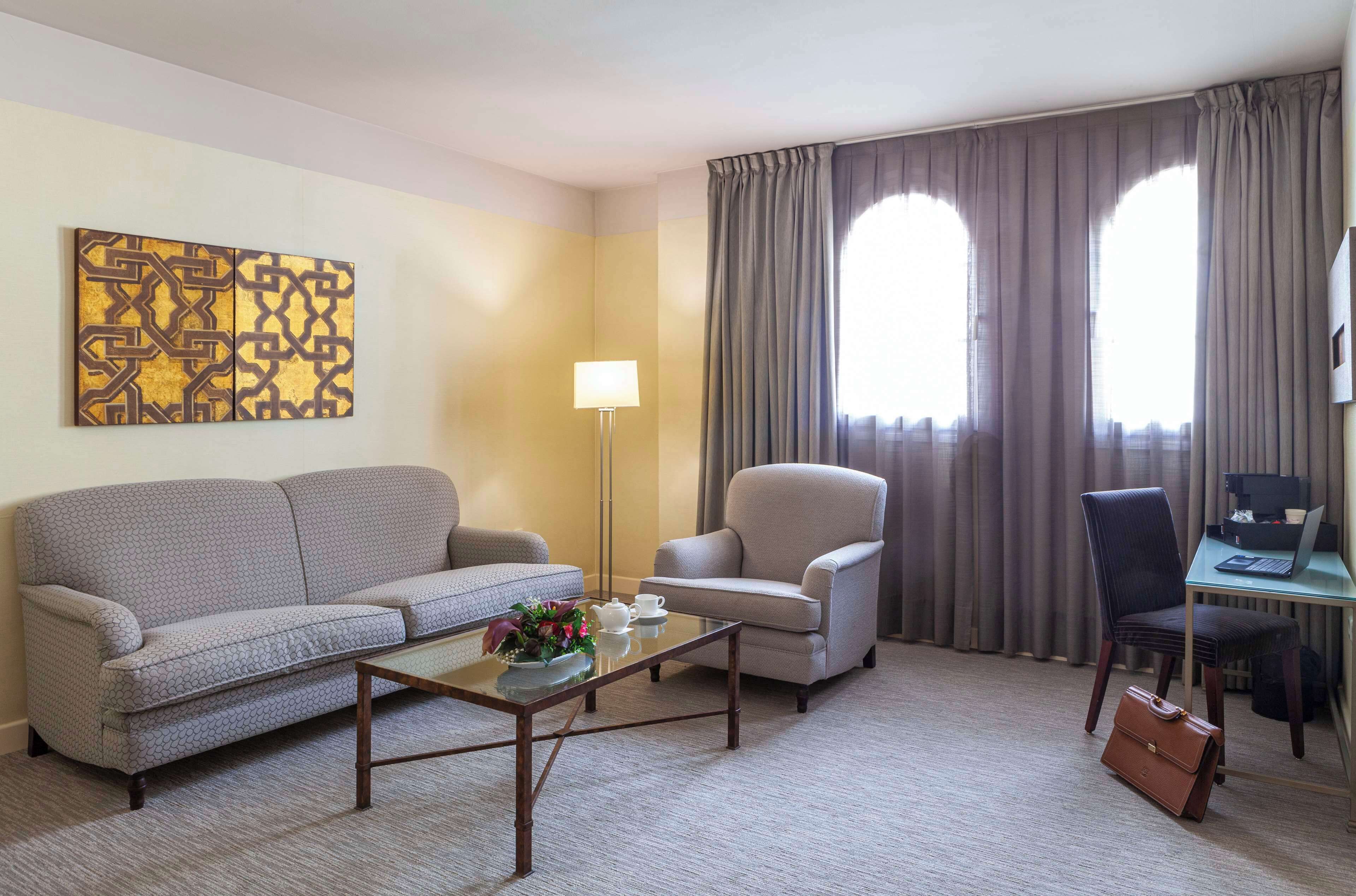 A hotel room with a sofa, armchair, desk, and a painting on the wall.