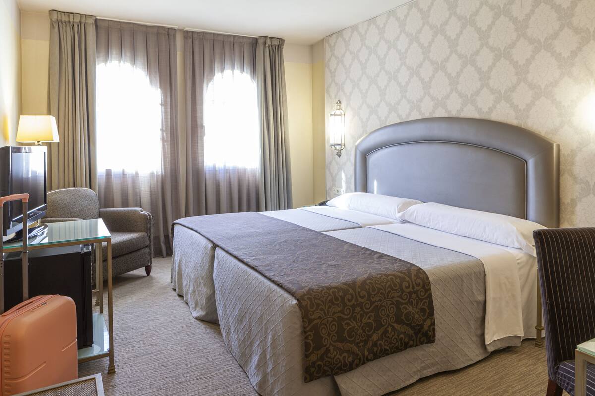 Ein Hotelzimmer mit einem großen Bett, einem Schreibtisch mit Computer, einem Sessel und einer Lampe.