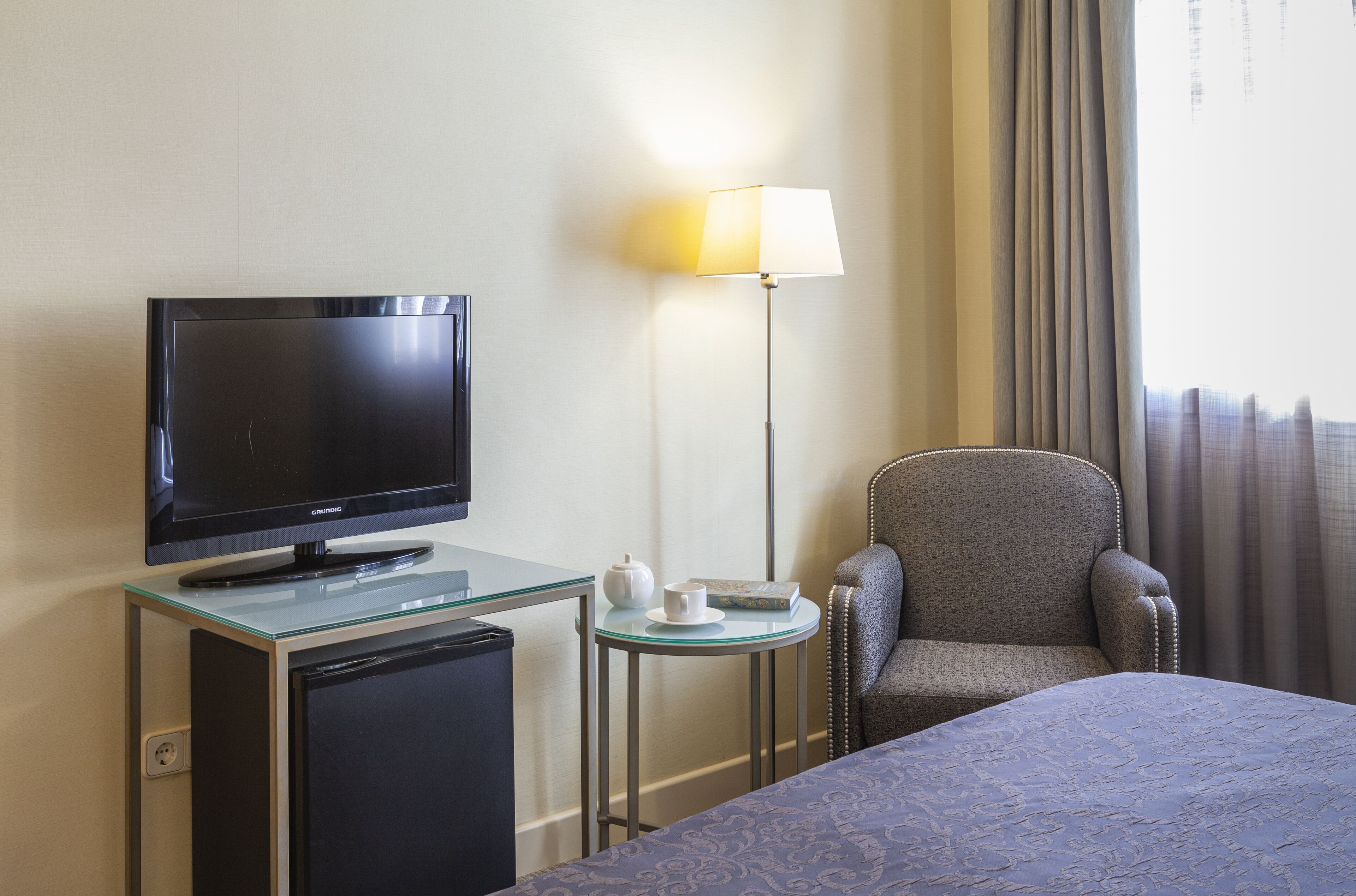 A hotel room with a television on a glass table, a small side table with a teapot and cup, a comfortable armchair, and a bed with a patterned bedspread.