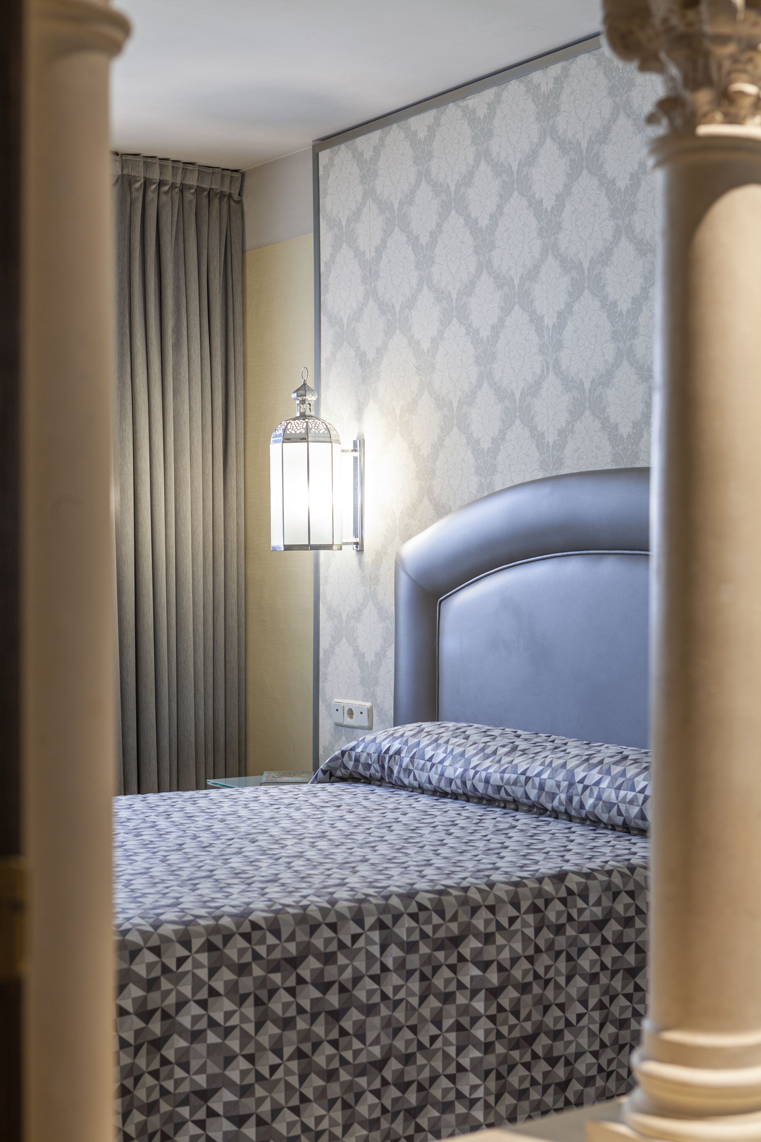Habitación de hotel con una cama, una lámpara fijada a la pared y un papel tapiz estampado.