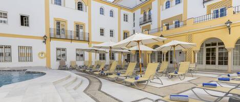 Zona de piscina con tumbonas y sombrillas, rodeada por un edificio con balcones y ventanas amplias.